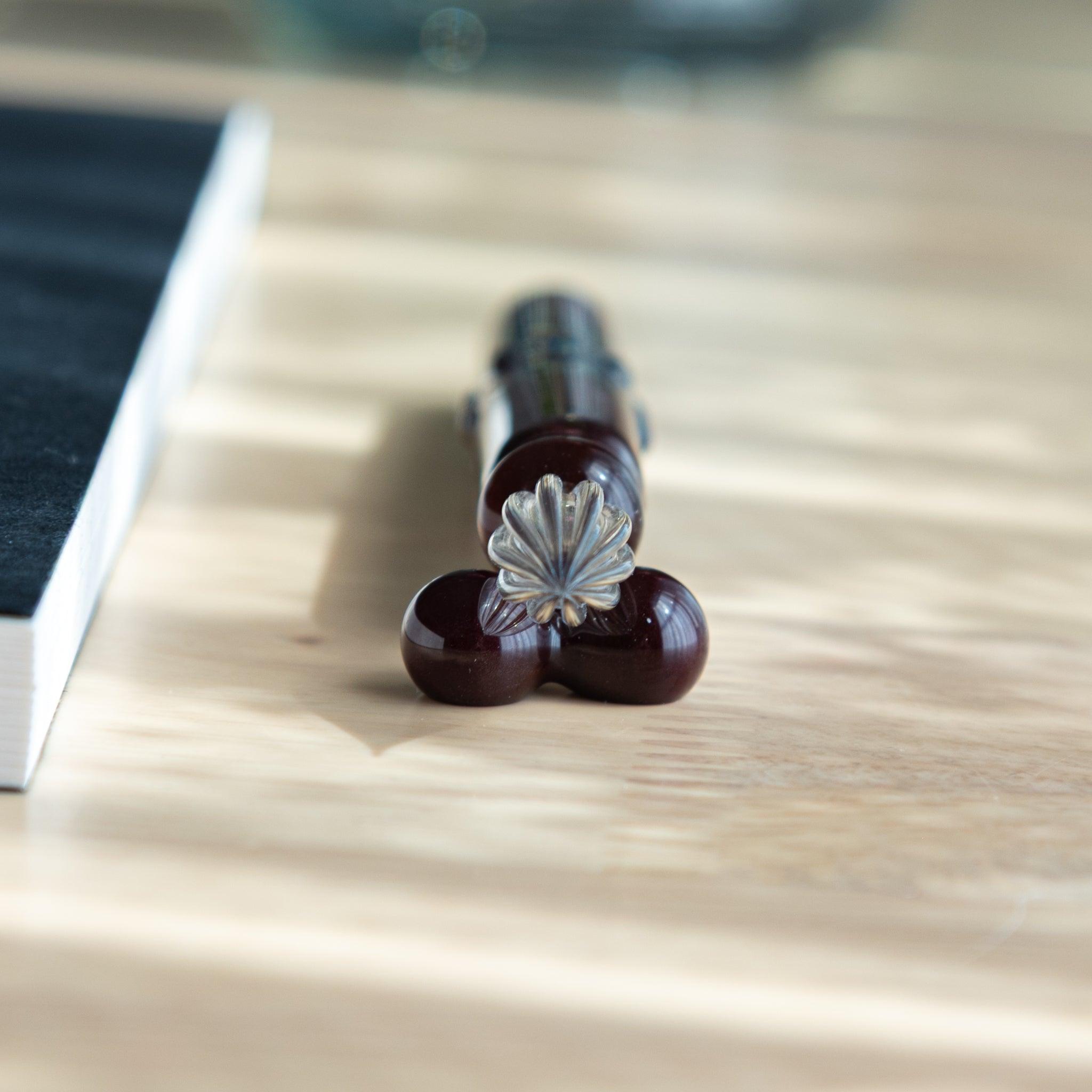 Front view of a glass dip pen handmade in Japan, showing its clear nib and dark maroon body supported by a pen rest.