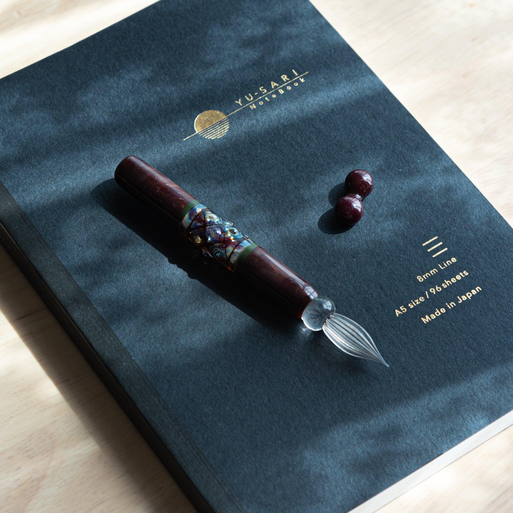 A maroon glass dip pen from Japan with a decorative band lies diagonally on a black notebook under natural light.