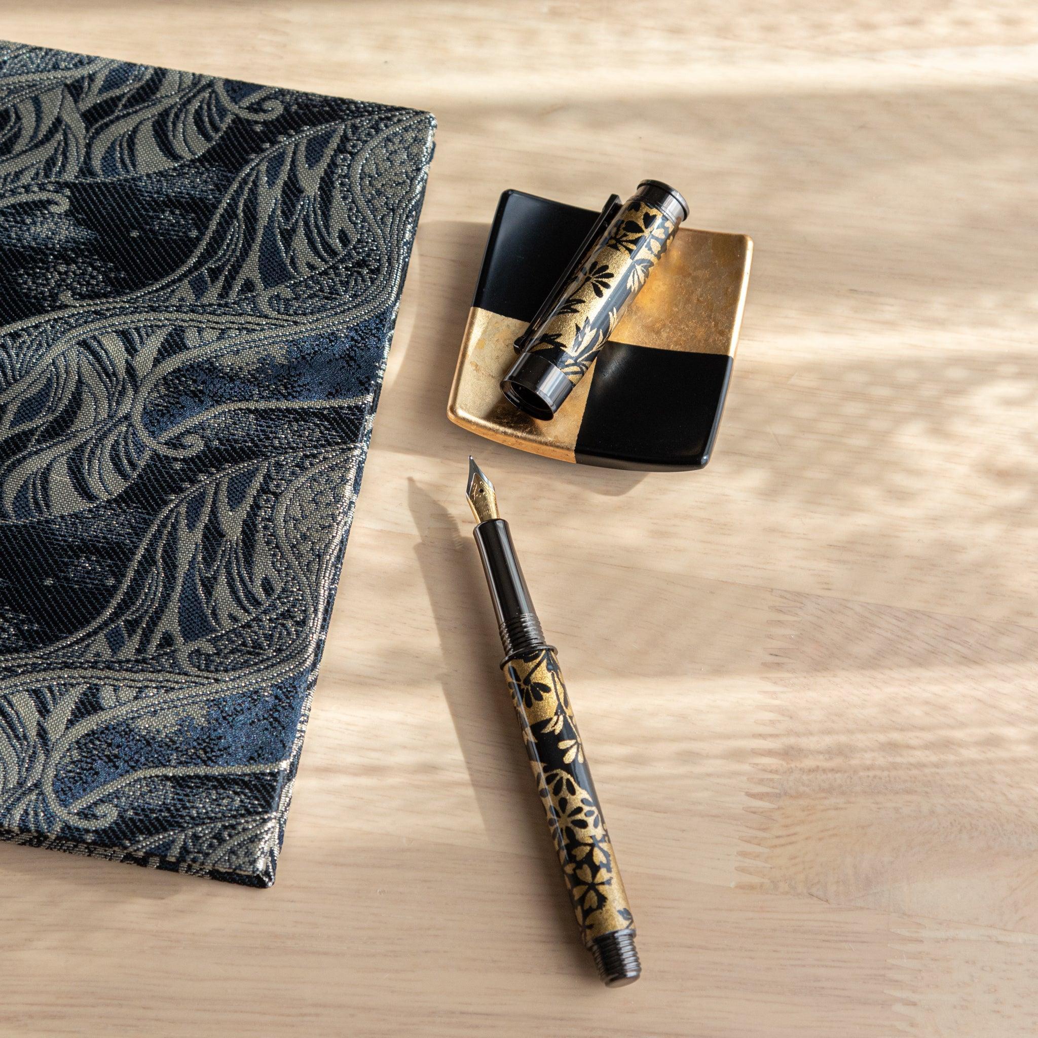 A Japanese stationery fountain pen with black and gold floral designs, disassembled, placed beside a patterned fabric and dish.