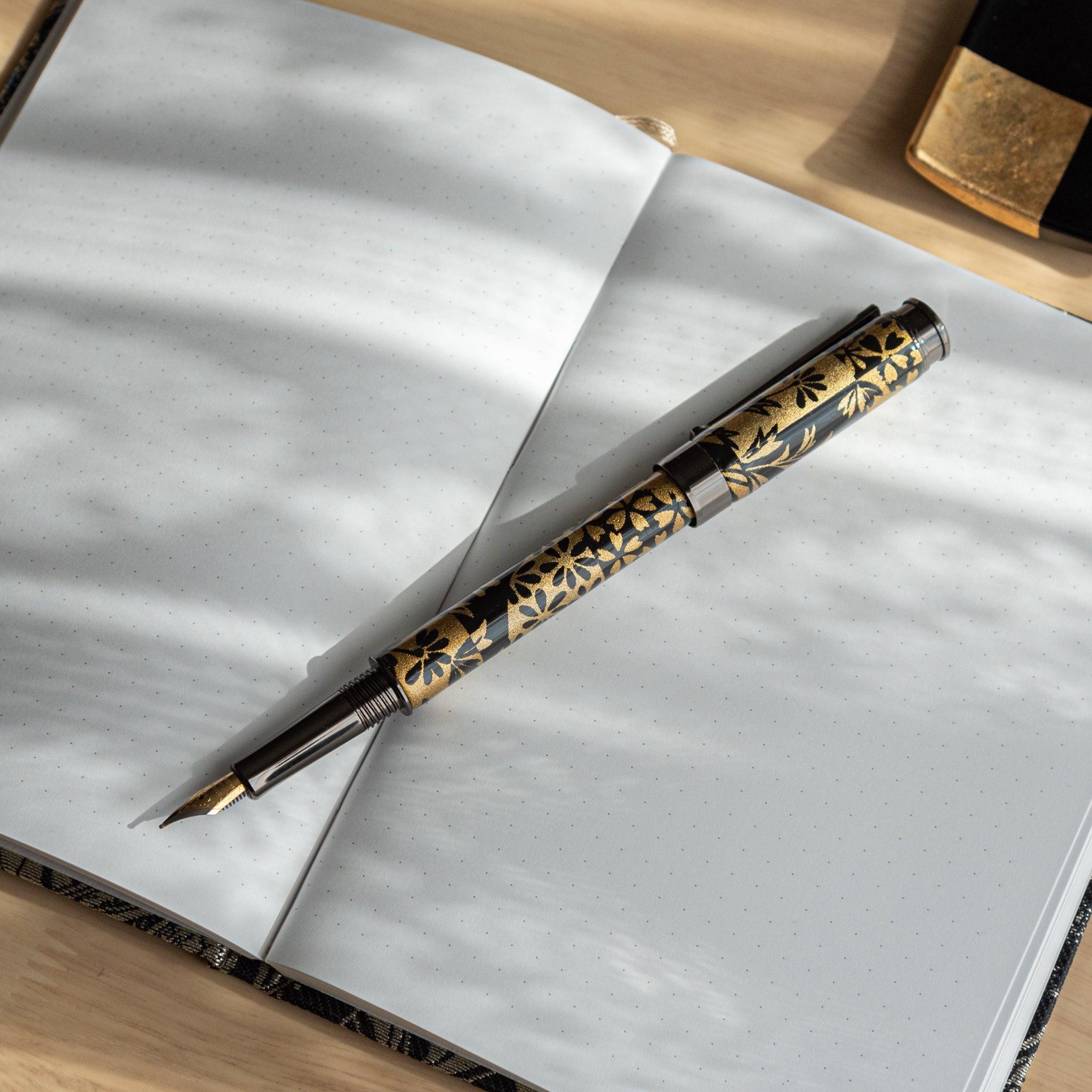 A Japanese stationery fountain pen with black and gold floral patterns, placed on an open dotted notebook under soft light.