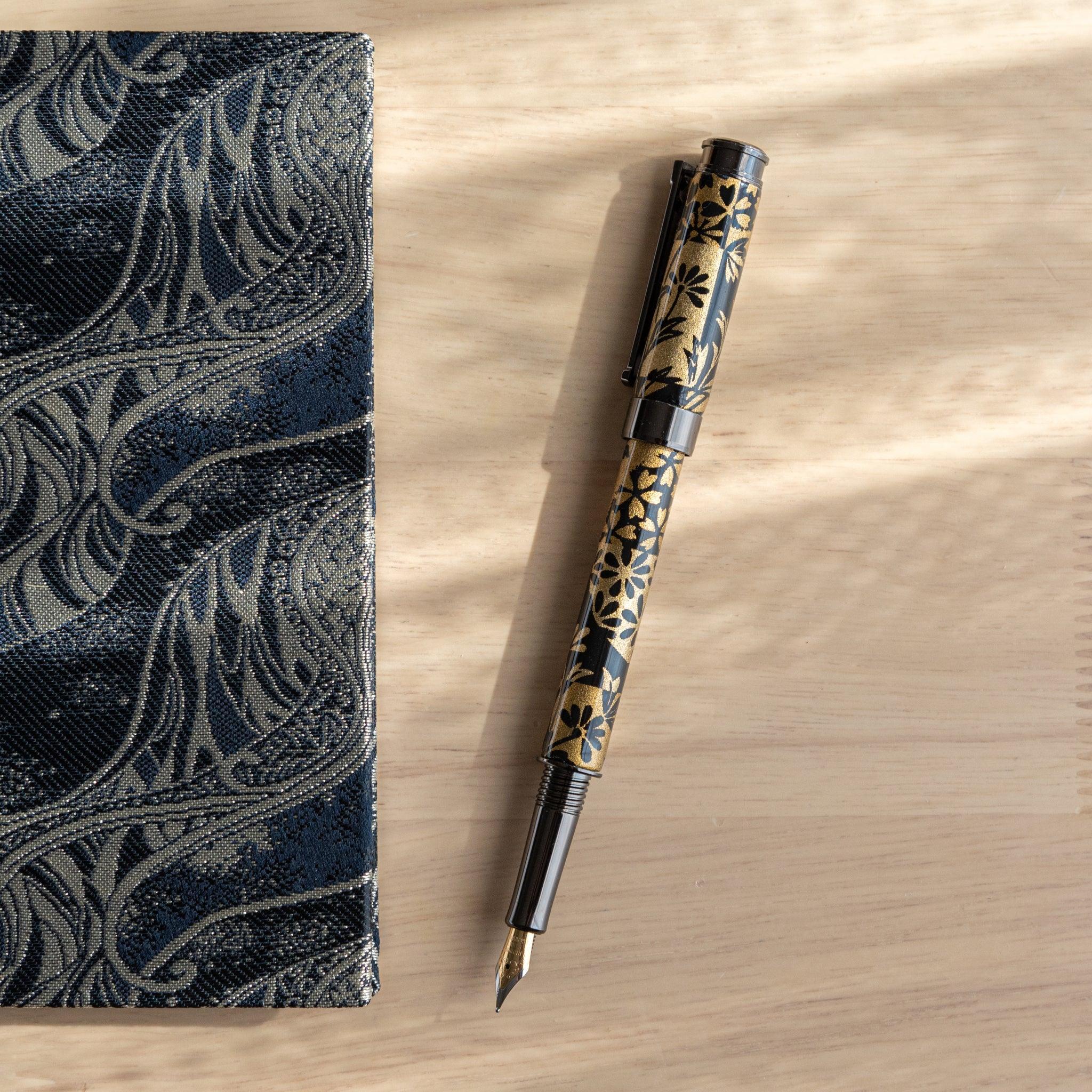 A Japanese stationery fountain pen with black and gold floral patterns, placed next to a patterned notebook on a wooden surface.