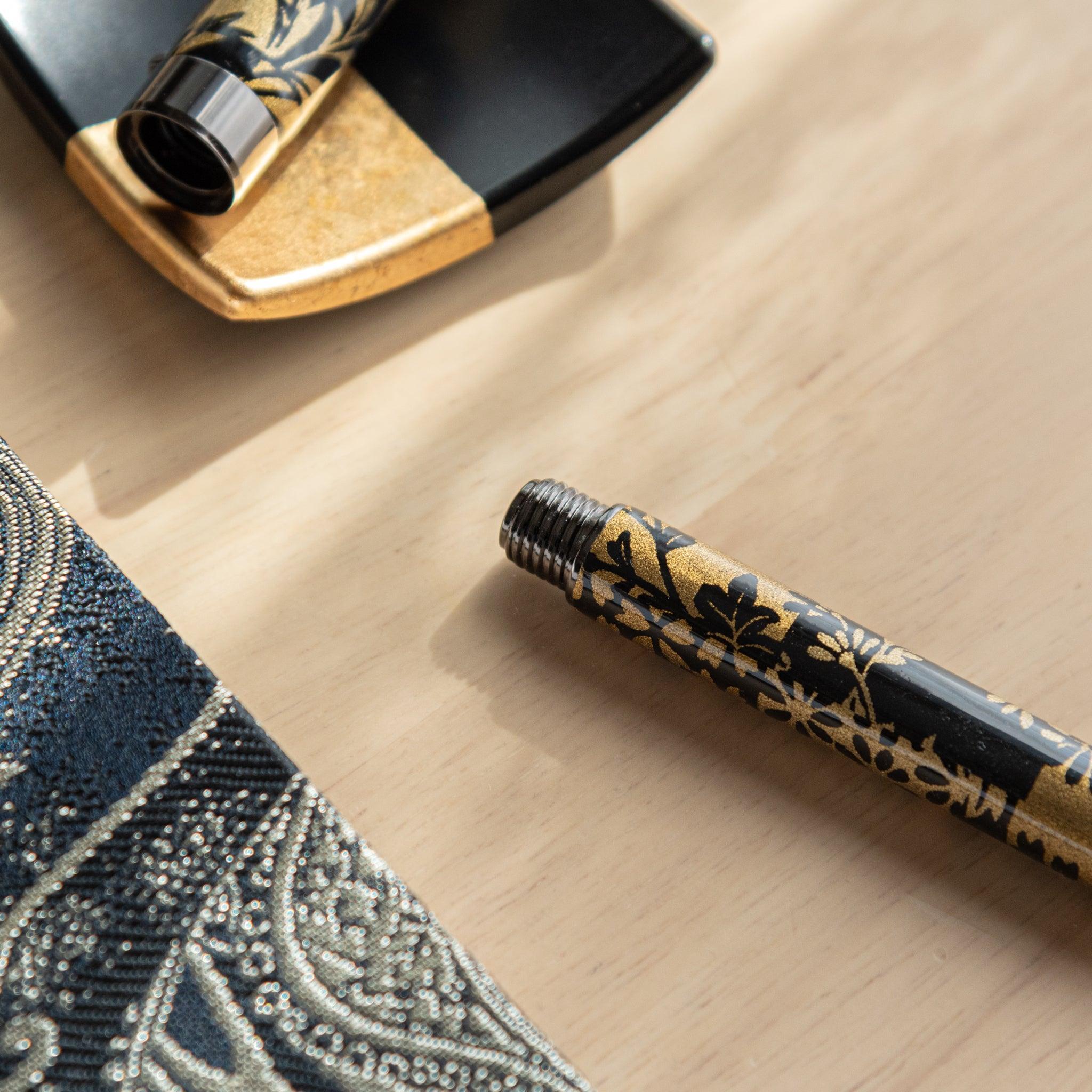 A Japanese stationery fountain pen barrel with black and gold floral details, highlighting the threaded end, near a decorative dish.