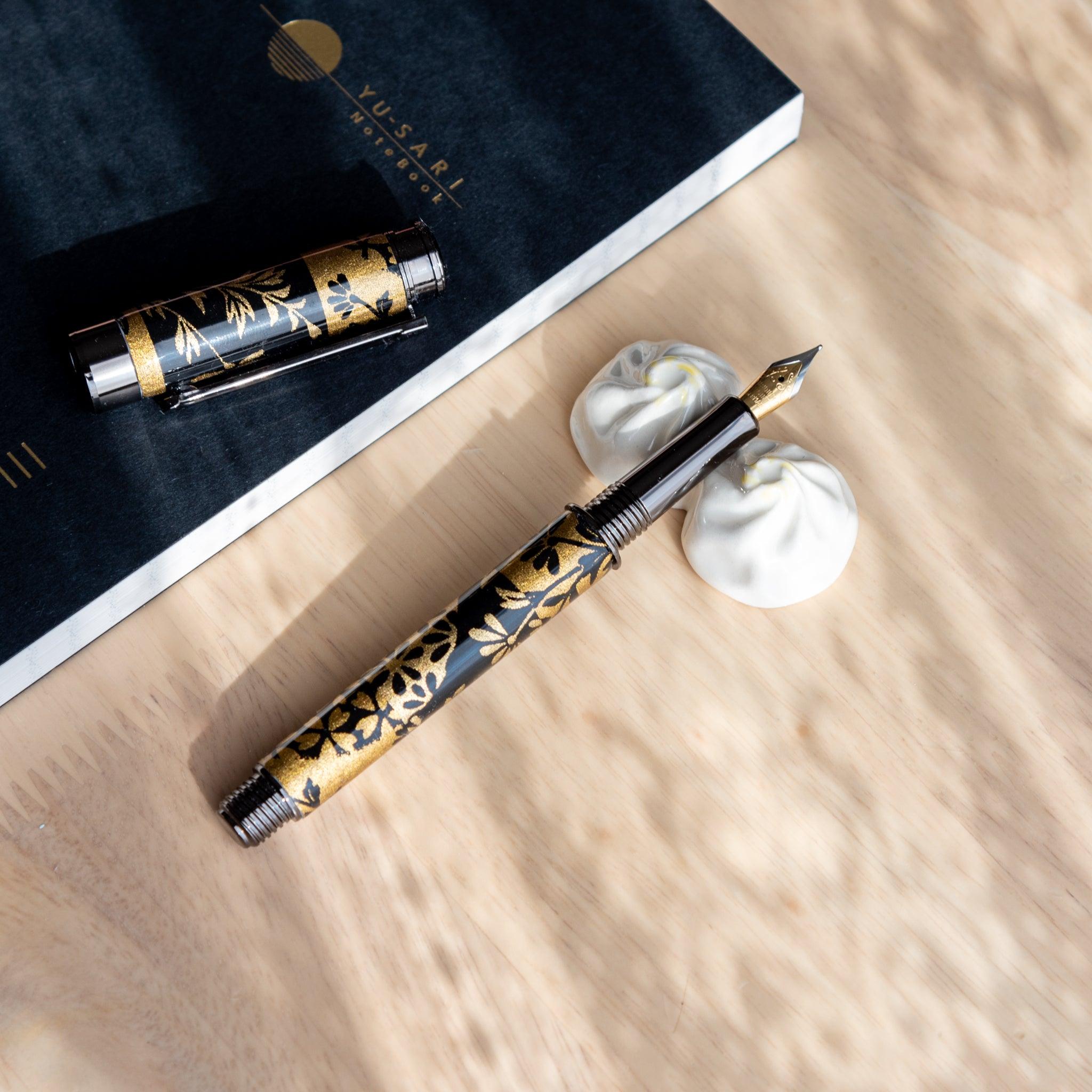 A Japanese fountain pen featuring Yuzen-patterned Mino washi rests on a white ceramic holder. A black Yusari notebook lies nearby on a wooden desk.