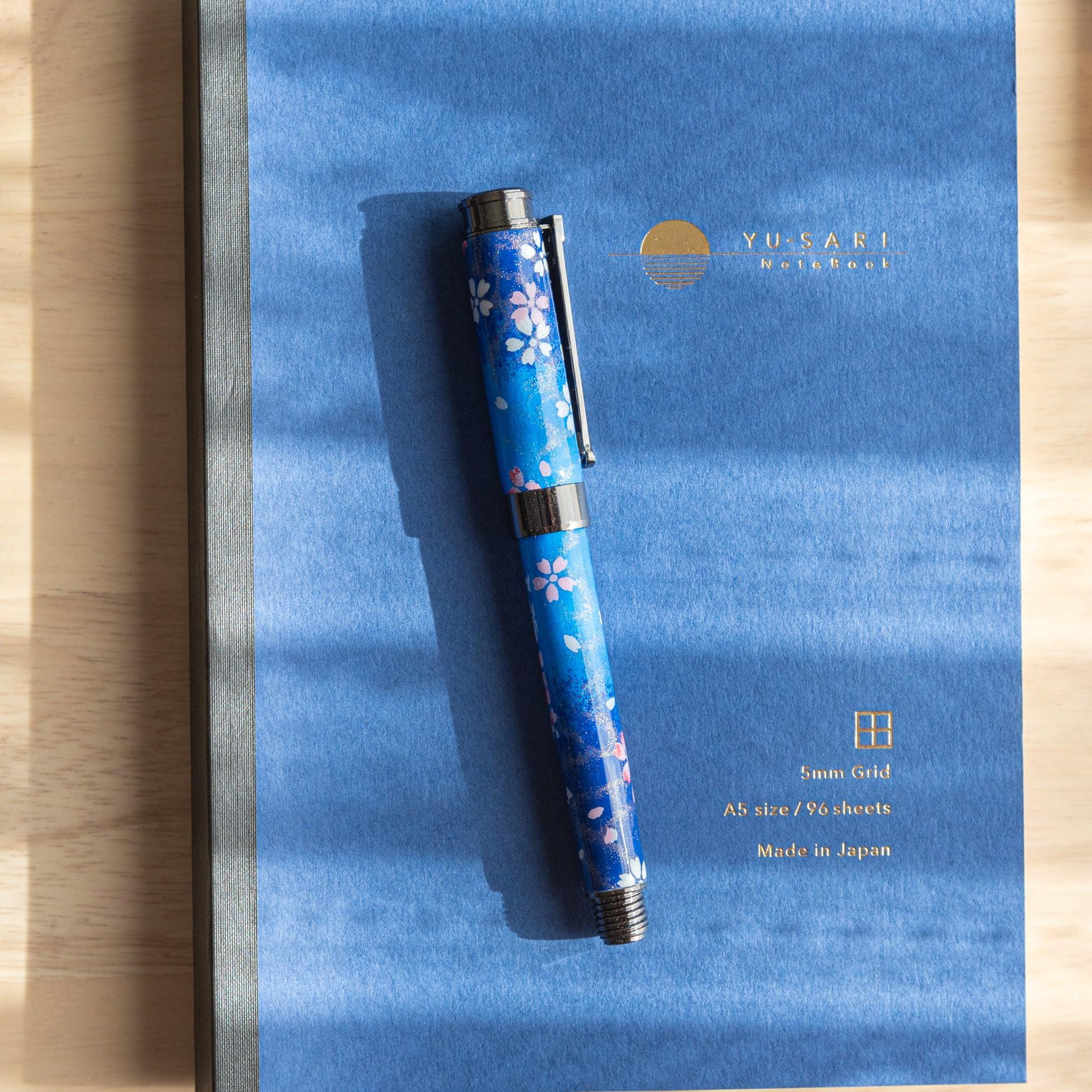 Japanese fountain pen with sakura motif in blue, placed on a blue notebook with gold lettering under soft light.