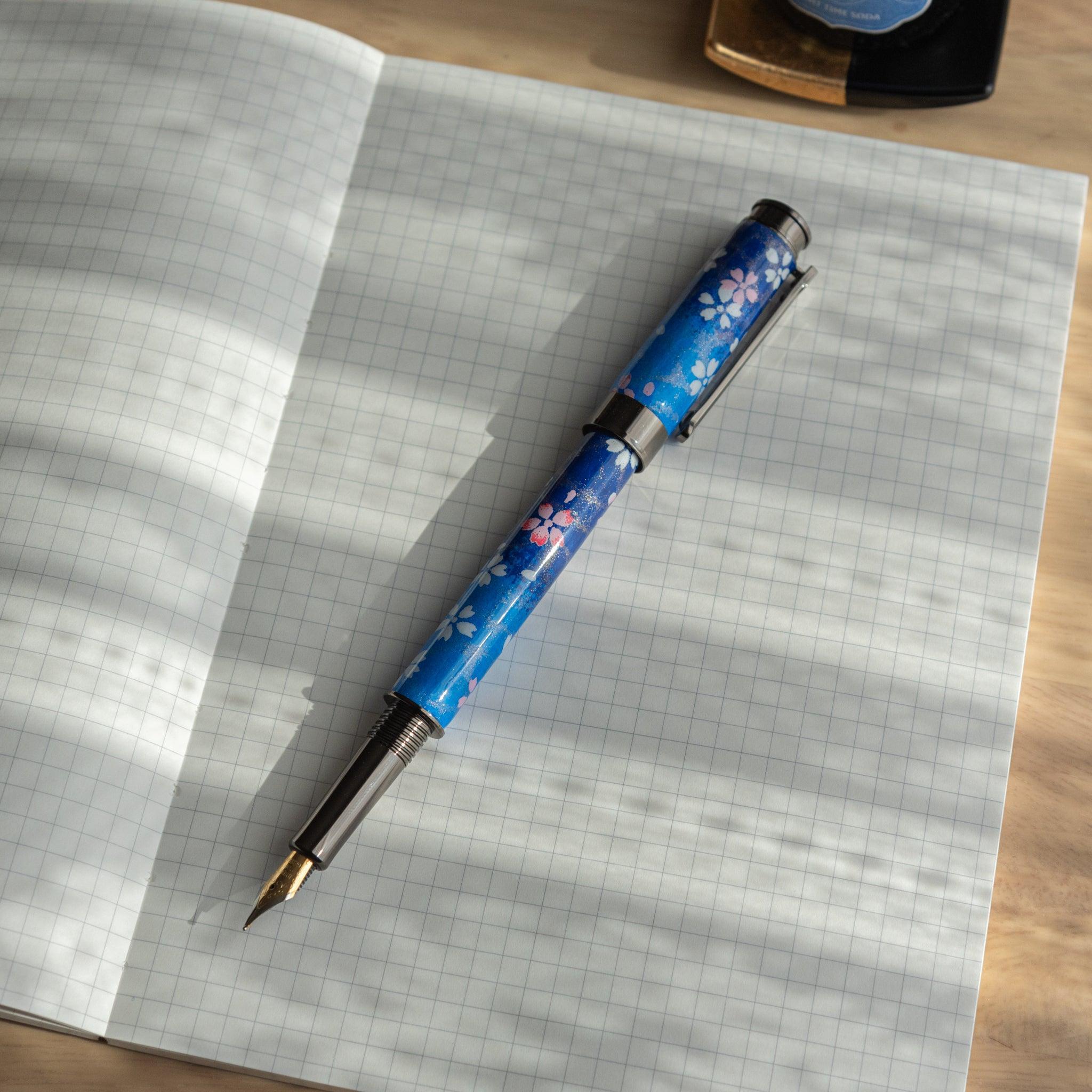 Japanese fountain pen with blue sakura pattern, open and placed on a grid-lined notebook under natural light.
