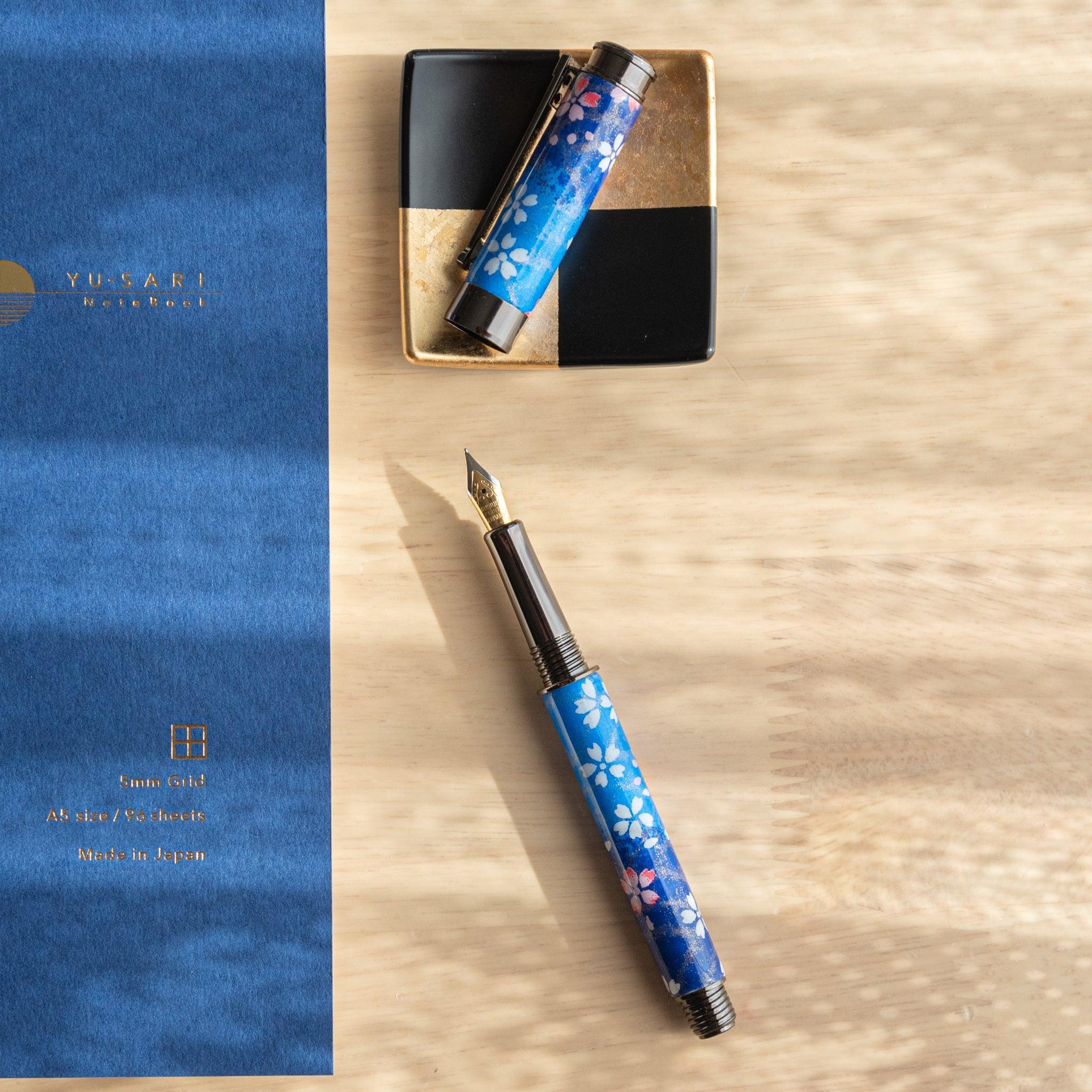 Open Japanese fountain pen with moonlit sakura motif in blue, displayed with cap on a gold-accented tray and notebook.