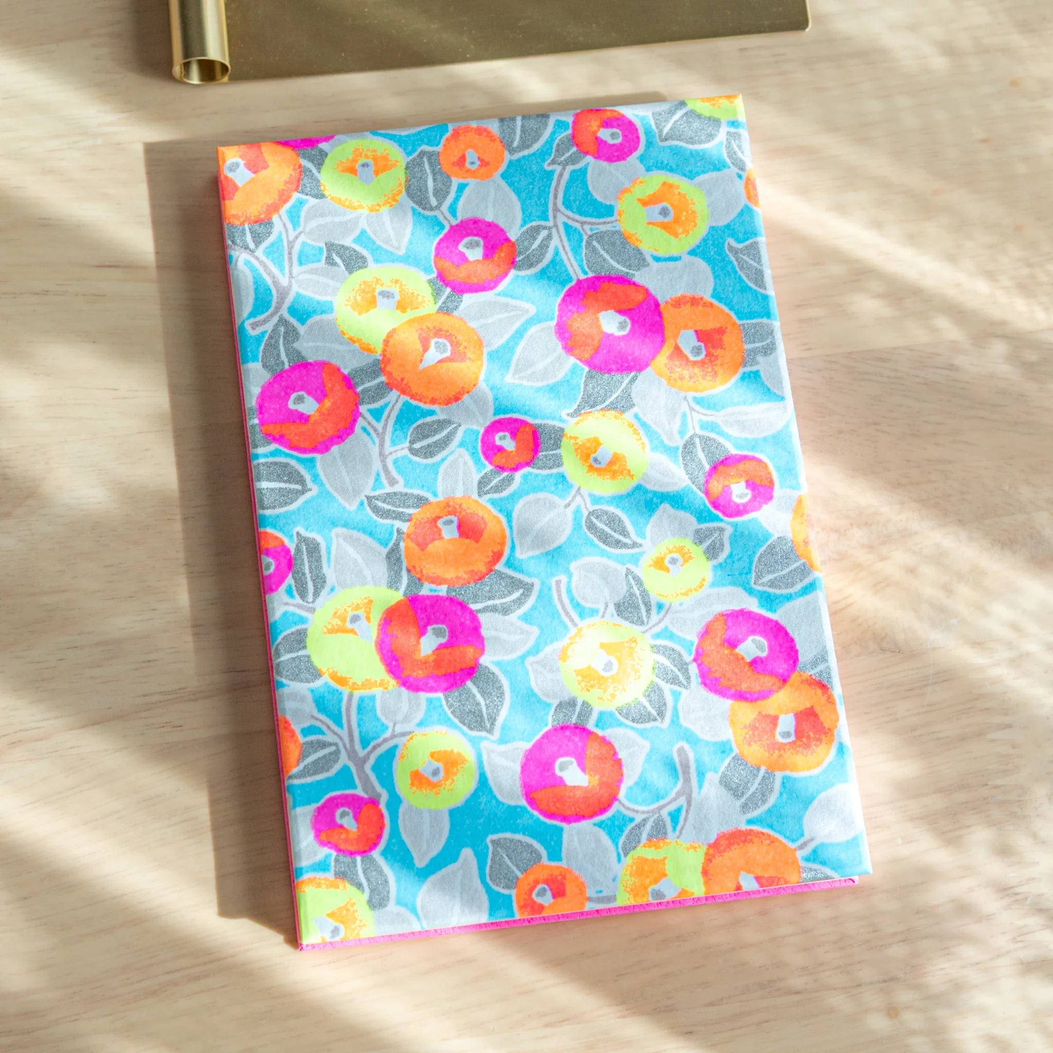 Neon Camellia washi goshuincho front cover in light blue with neon multicolor camellia blooms and grey leaves on light wood