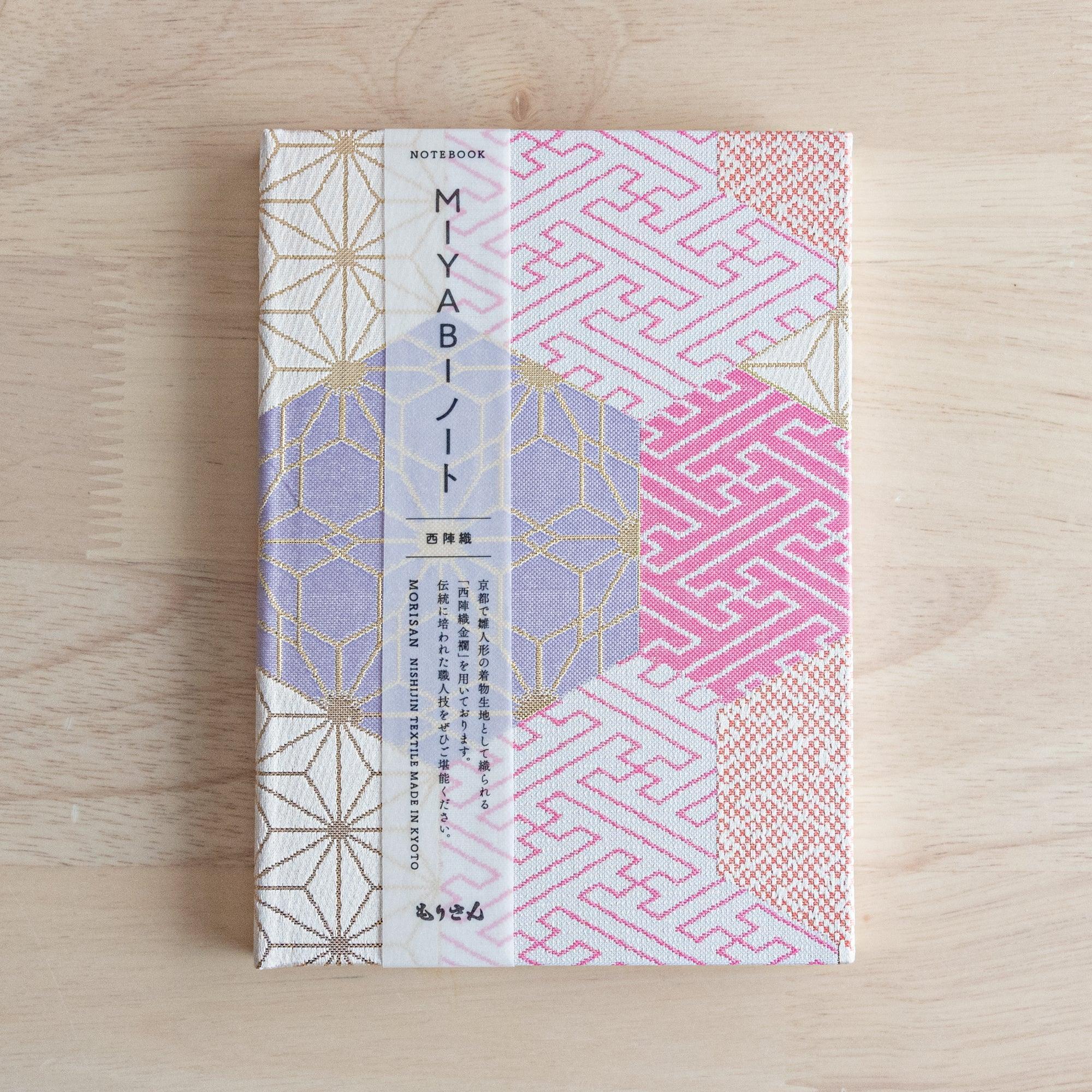 A Japanese notebook with a fabric cover featuring traditional geometric patterns in pink, lavender, and gold tones, placed on a wooden table.