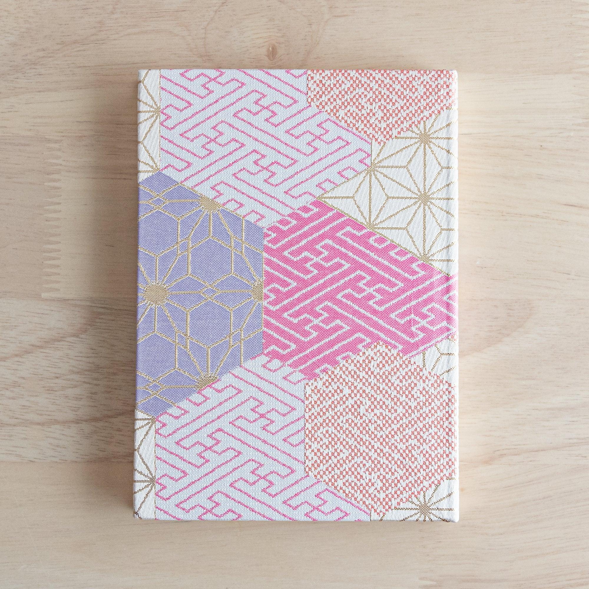A Japanese notebook with a fabric cover featuring traditional geometric patterns in pink, lavender, and gold tones, placed on a wooden table.