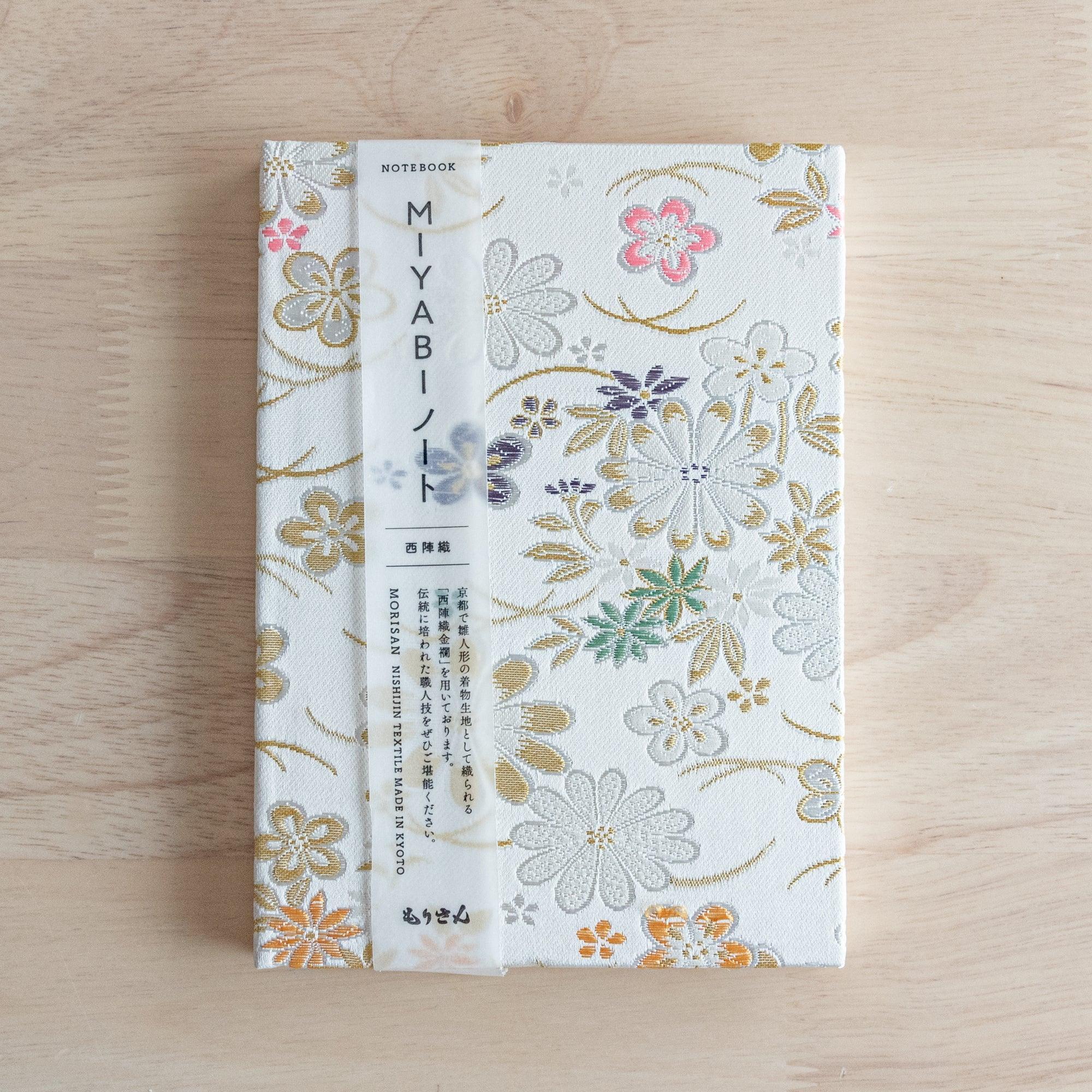 A Japanese notebook with a white fabric cover adorned with colorful floral embroidery in gold, green, purple, and pink, placed on a wooden table.