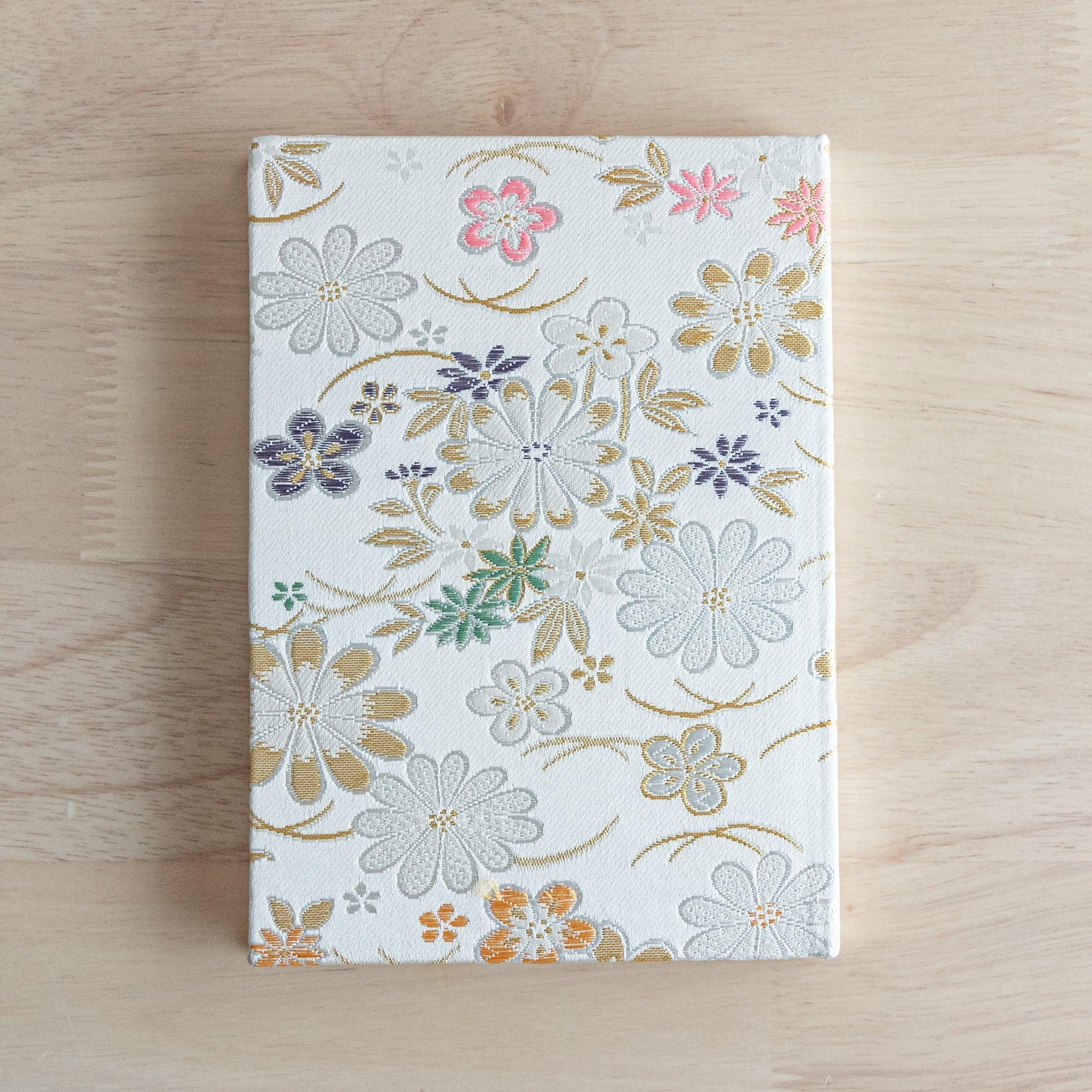 A Japanese notebook with a white fabric cover adorned with colorful floral embroidery in gold, green, purple, and pink, placed on a wooden table.