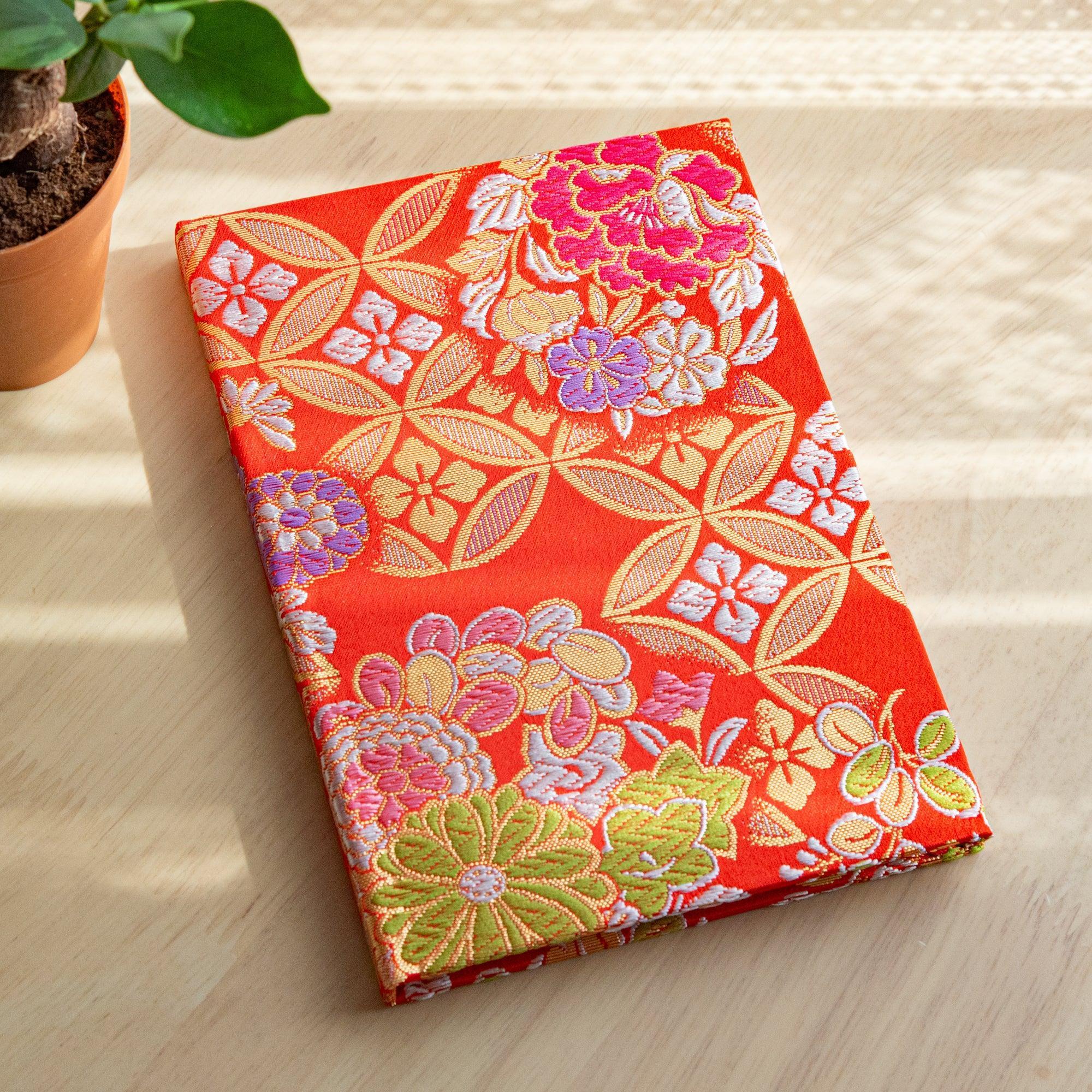 A Japanese notebook with a vibrant red fabric cover featuring intricate floral and geometric patterns in gold, pink, green, and purple.