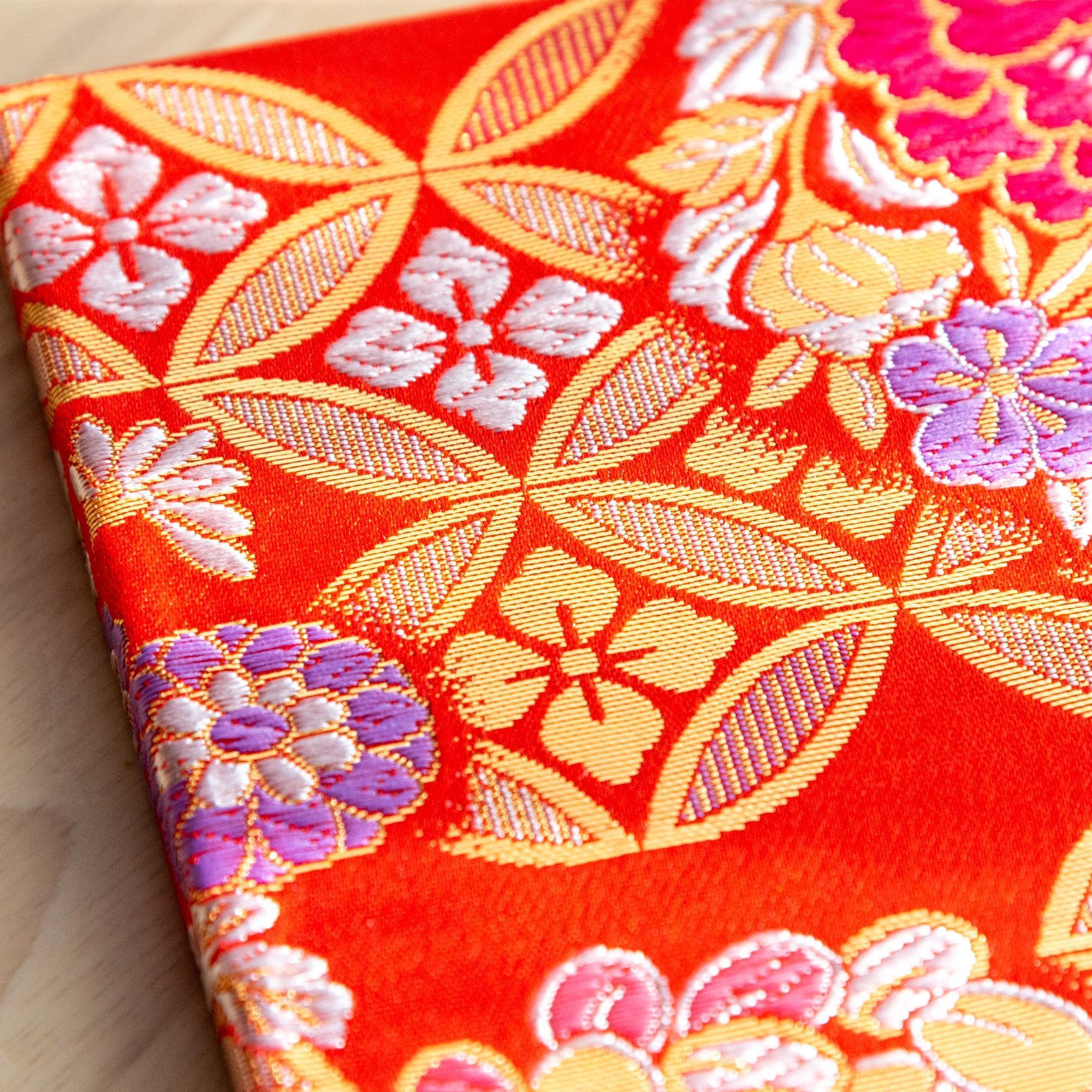 A Japanese notebook with a vibrant red fabric cover featuring intricate floral and geometric patterns in gold, pink, green, and purple.