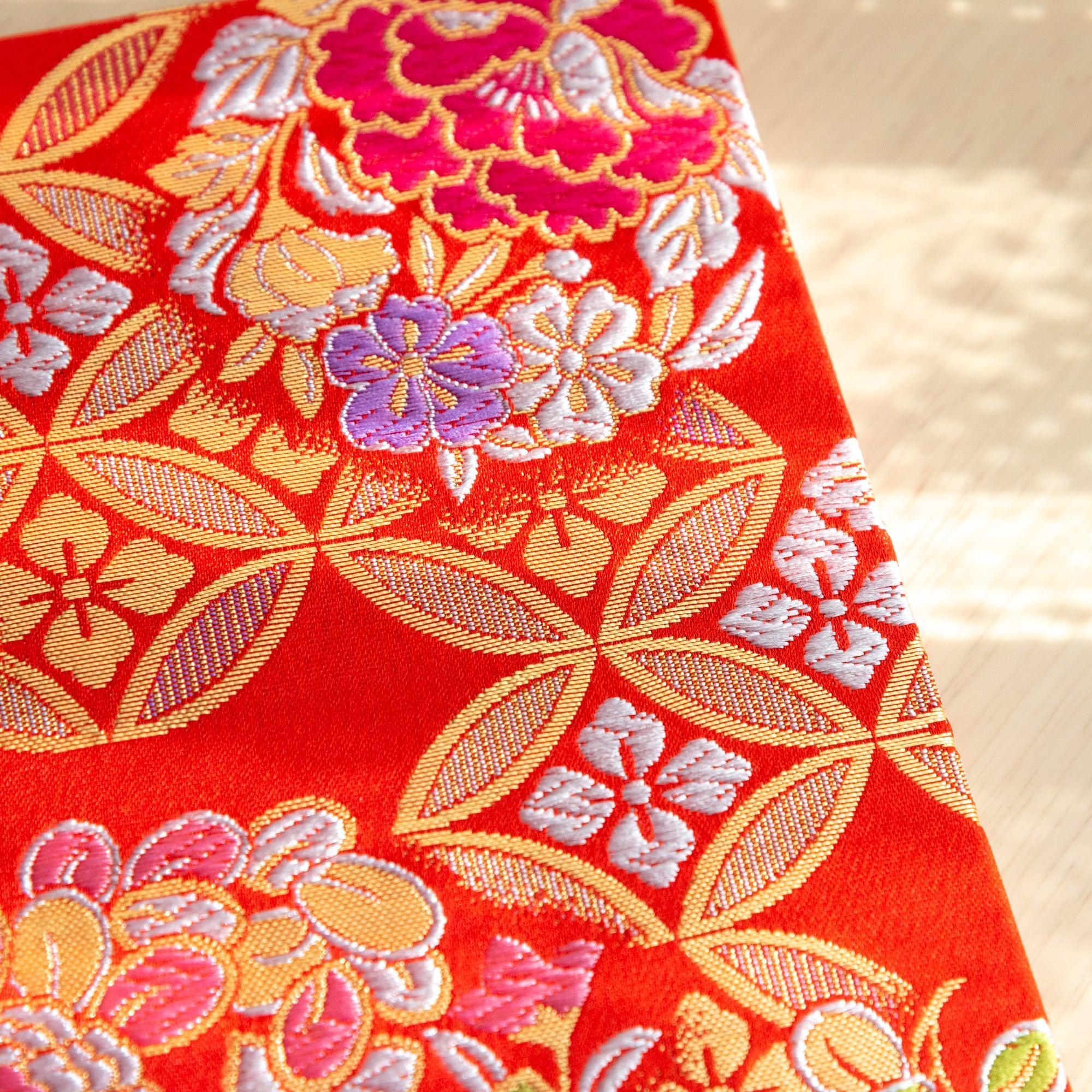 A Japanese notebook with a vibrant red fabric cover featuring intricate floral and geometric patterns in gold, pink, green, and purple.