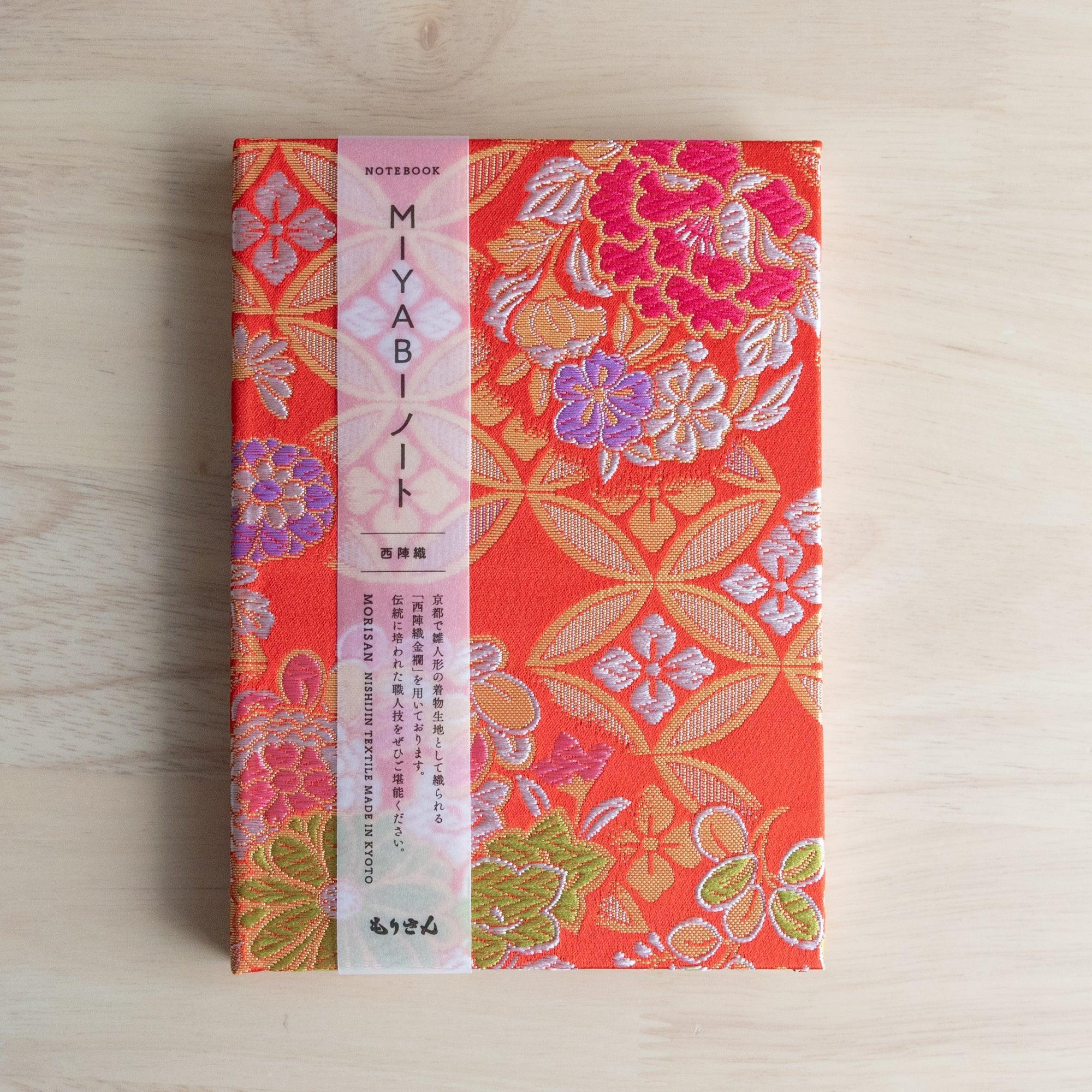 A Japanese notebook with a vibrant red fabric cover featuring intricate floral and geometric patterns in gold, pink, green, and purple.