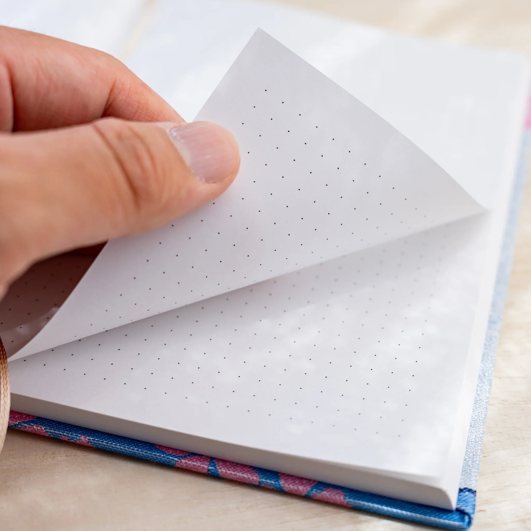 Hand turning a dot grid page of the notebook, showing paper thickness and blue and pink textile cover