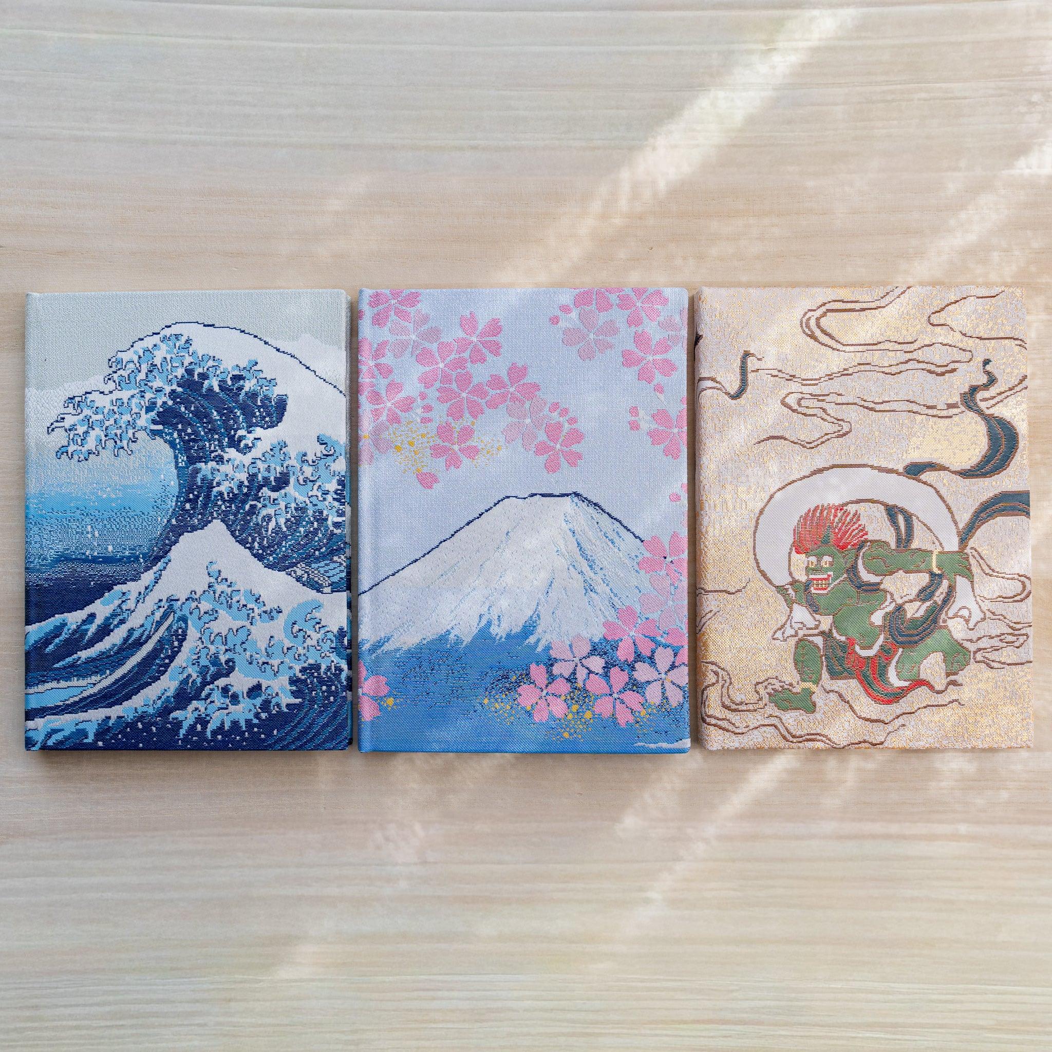 Three notebook covers side by side in woven textile: Great Wave, Mount Fuji with cherry blossoms, and Fujin Raijin gods