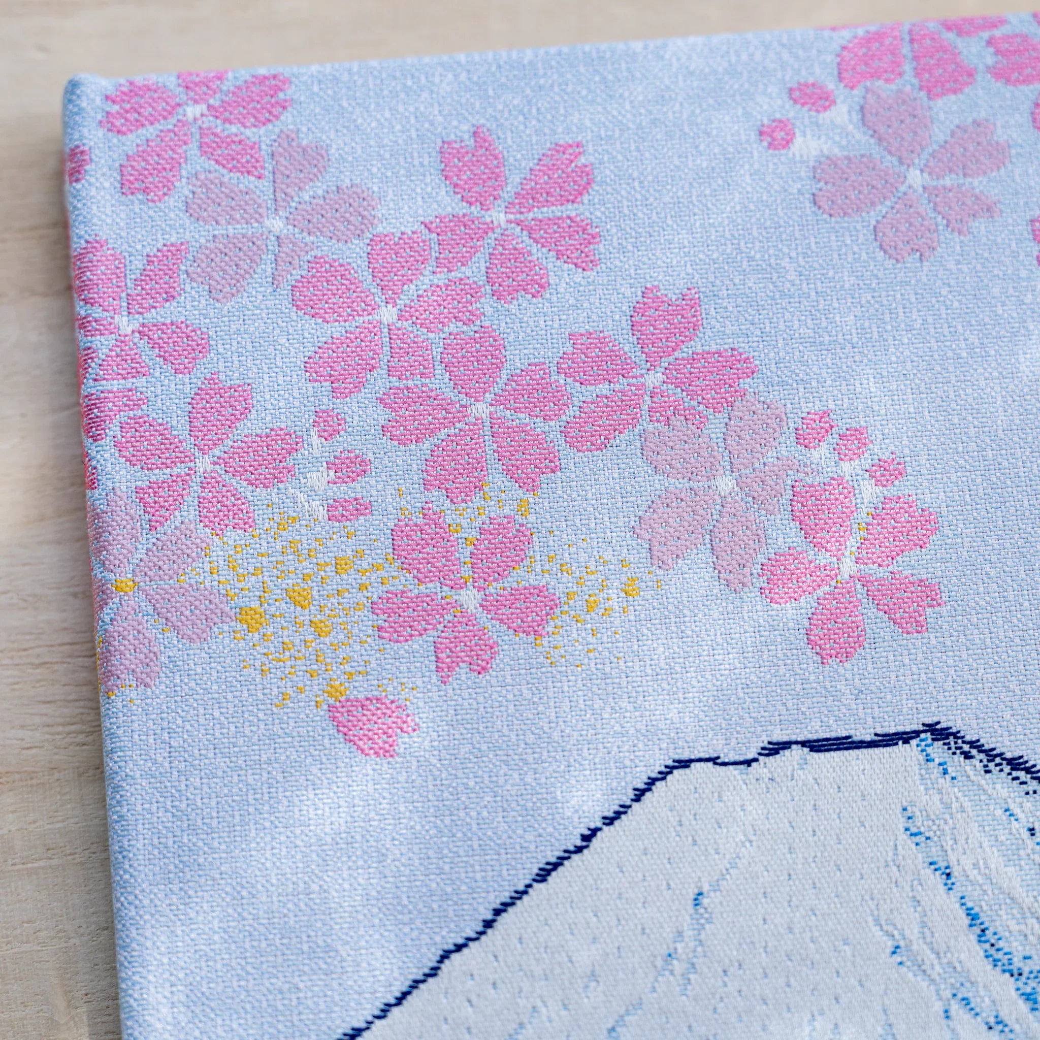 Close-up of woven textile showing pink cherry blossoms with gold flecks on pale blue, edge of Mount Fuji below