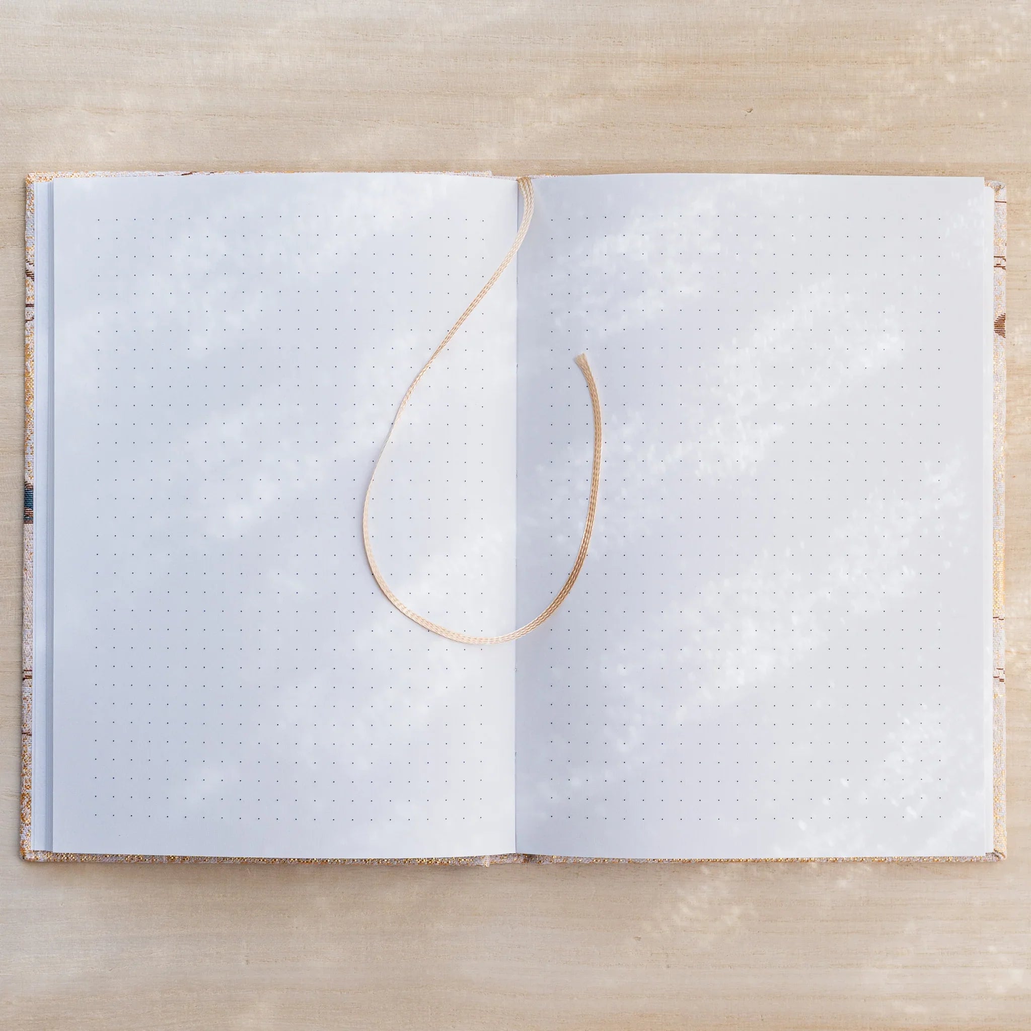 Notebook open flat to dot grid pages on both sides with a beige ribbon bookmark, gold textile cover edges