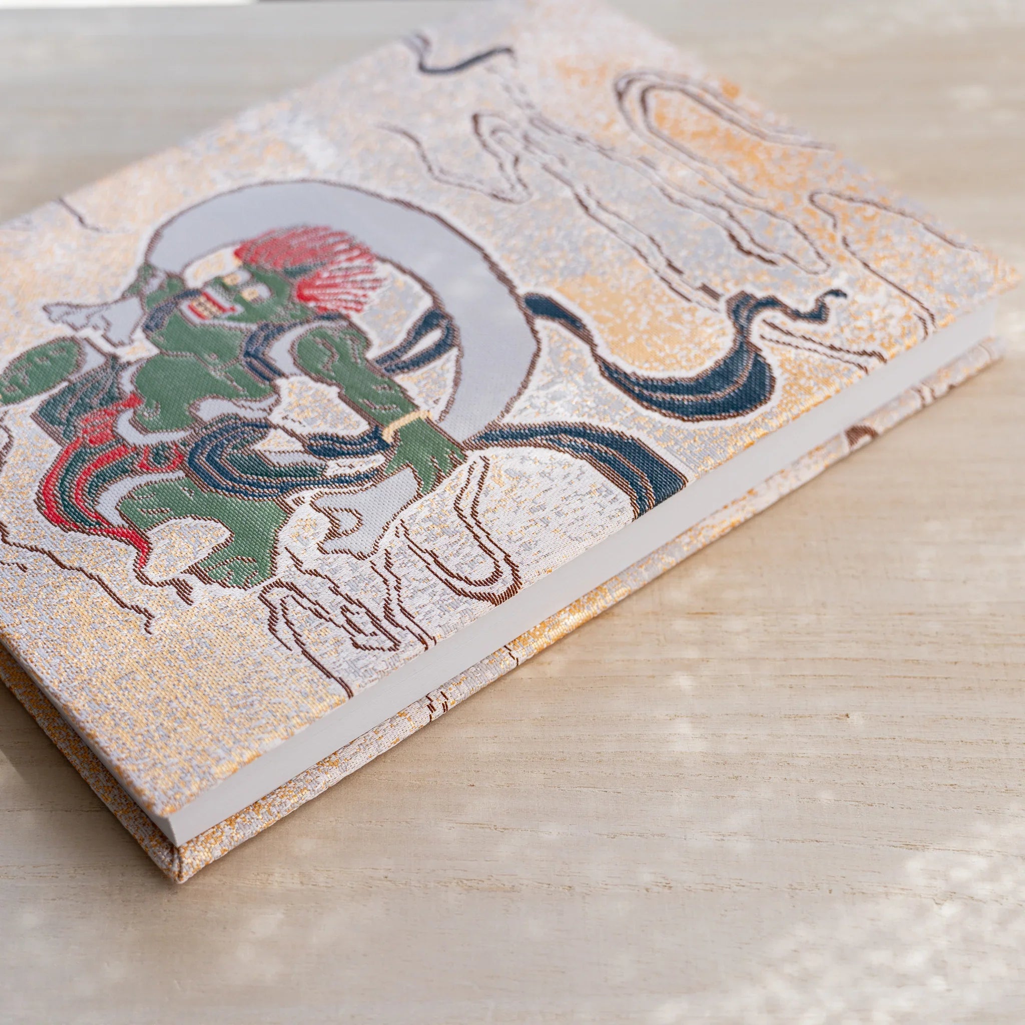 Angled view of notebook front cover, green wind god Fujin in woven textile on gold ground