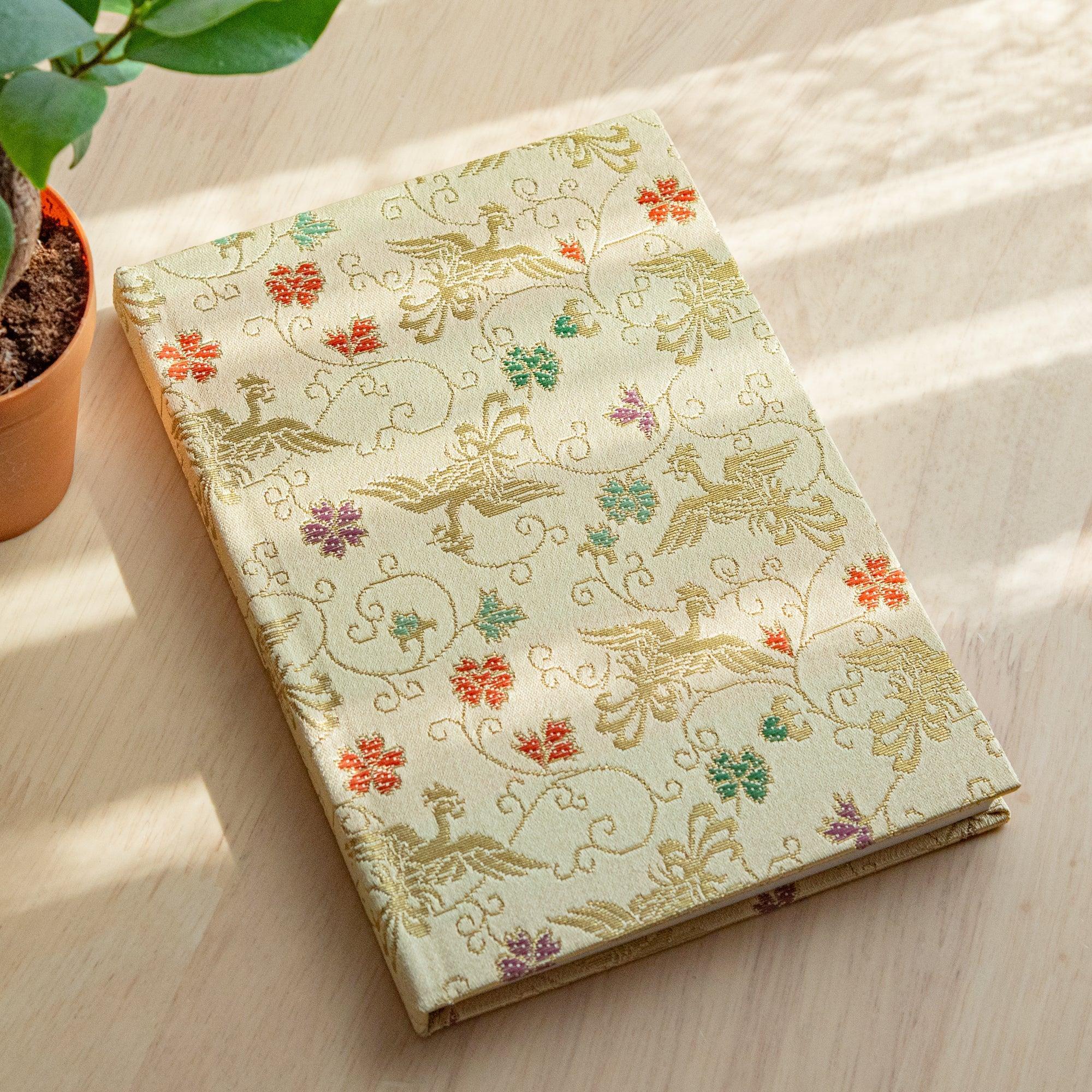 A Japanese notebook with an elegant gold fabric cover featuring intricate bird and floral patterns in red, green, and purple accents.