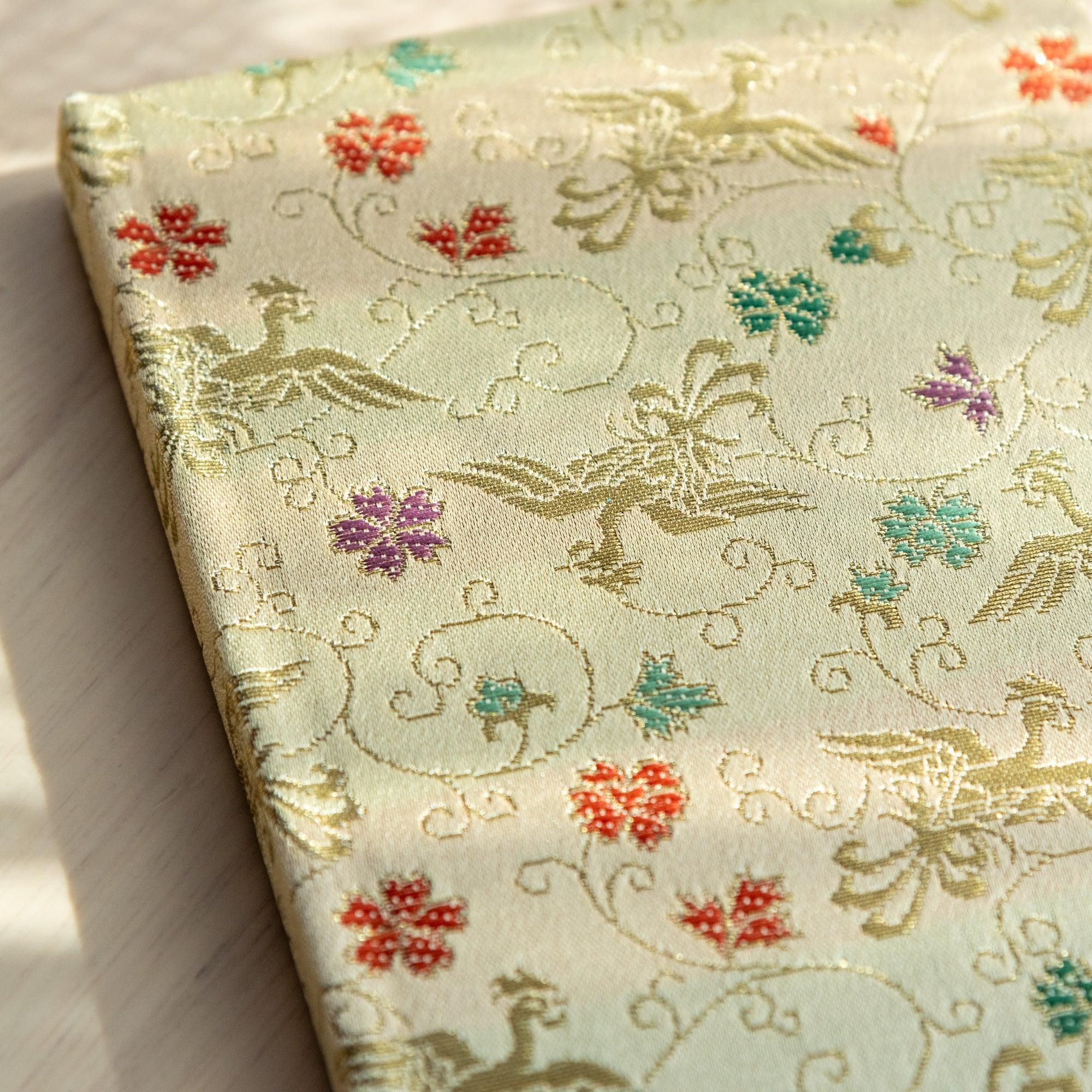 A Japanese notebook with an elegant gold fabric cover featuring intricate bird and floral patterns in red, green, and purple accents.