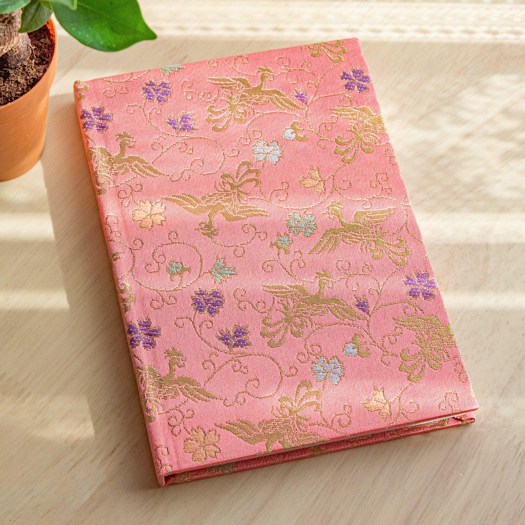 A Japanese notebook with a soft pink fabric cover featuring intricate gold bird and floral patterns accented with purple and green details.