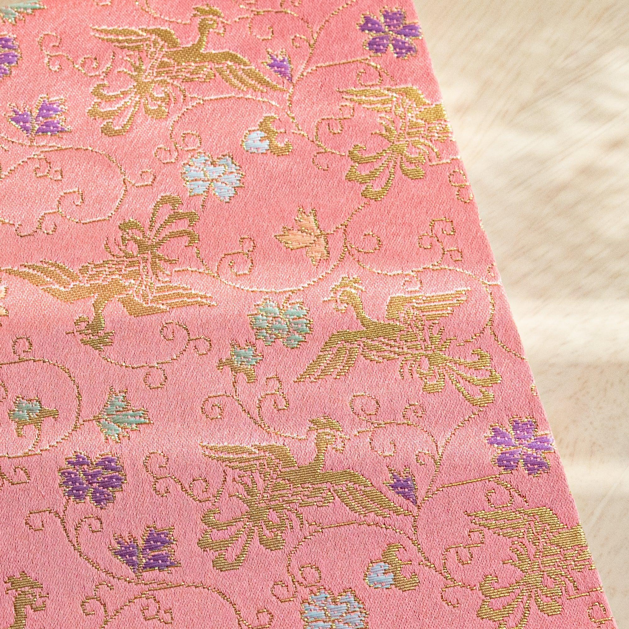 A Japanese notebook with a soft pink fabric cover featuring intricate gold bird and floral patterns accented with purple and green details.