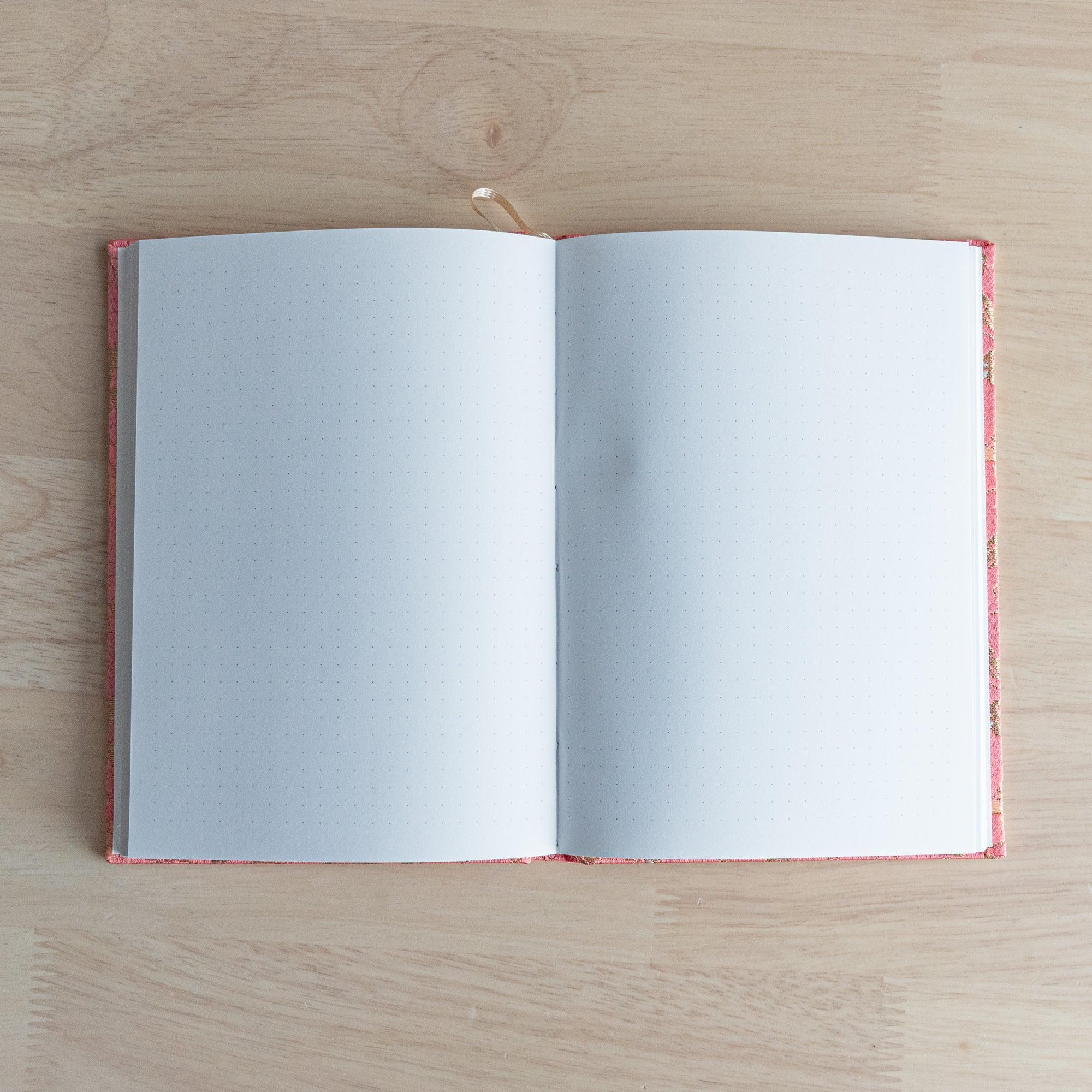 Opened Nishijin textile Japanese notebook showing two blank dot grid pages with pink woven textile cover and ribbon bookmark