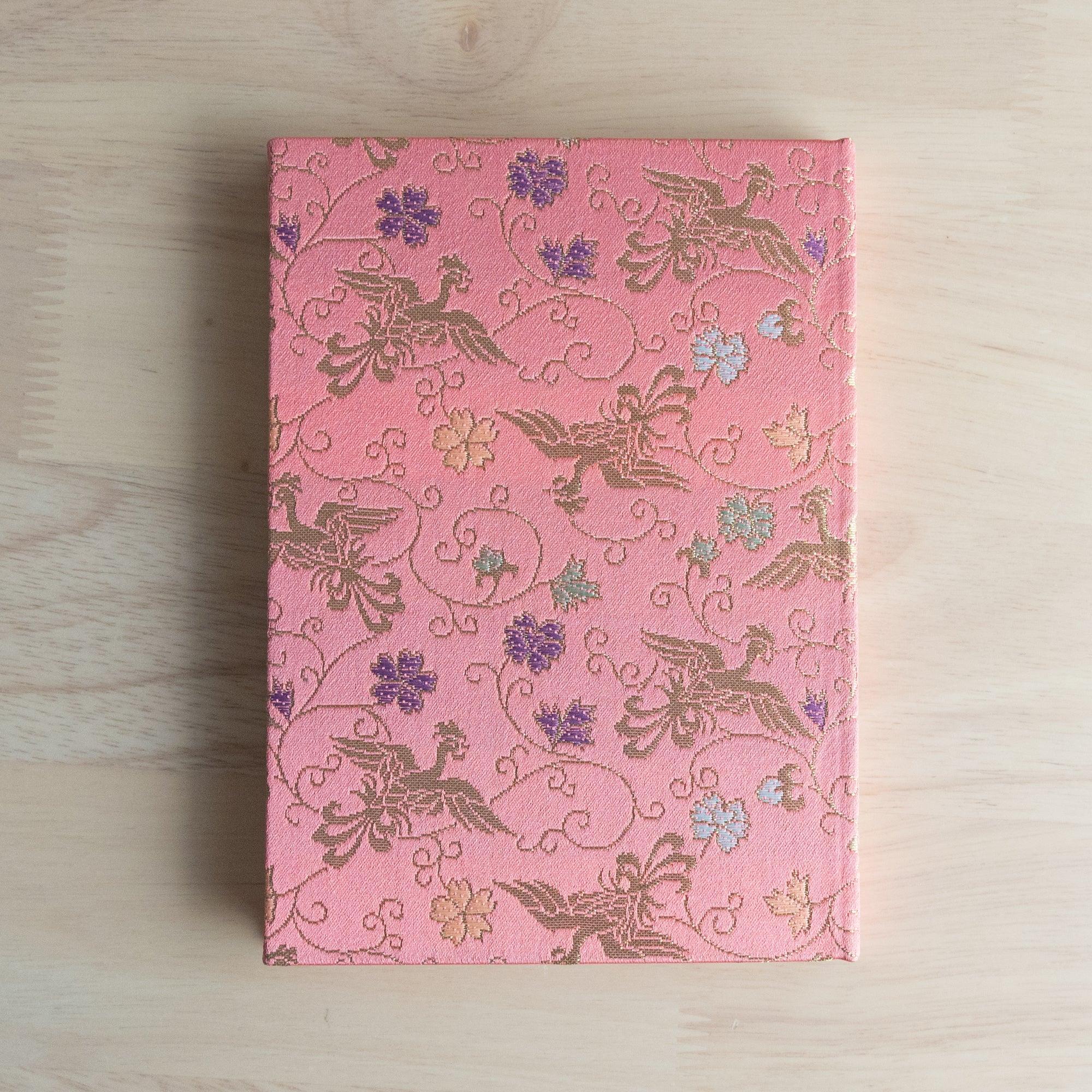 Japanese notebook with pink Nishijin woven textile cover showing gold phoenix birds and arabesque scrollwork, flat on wood