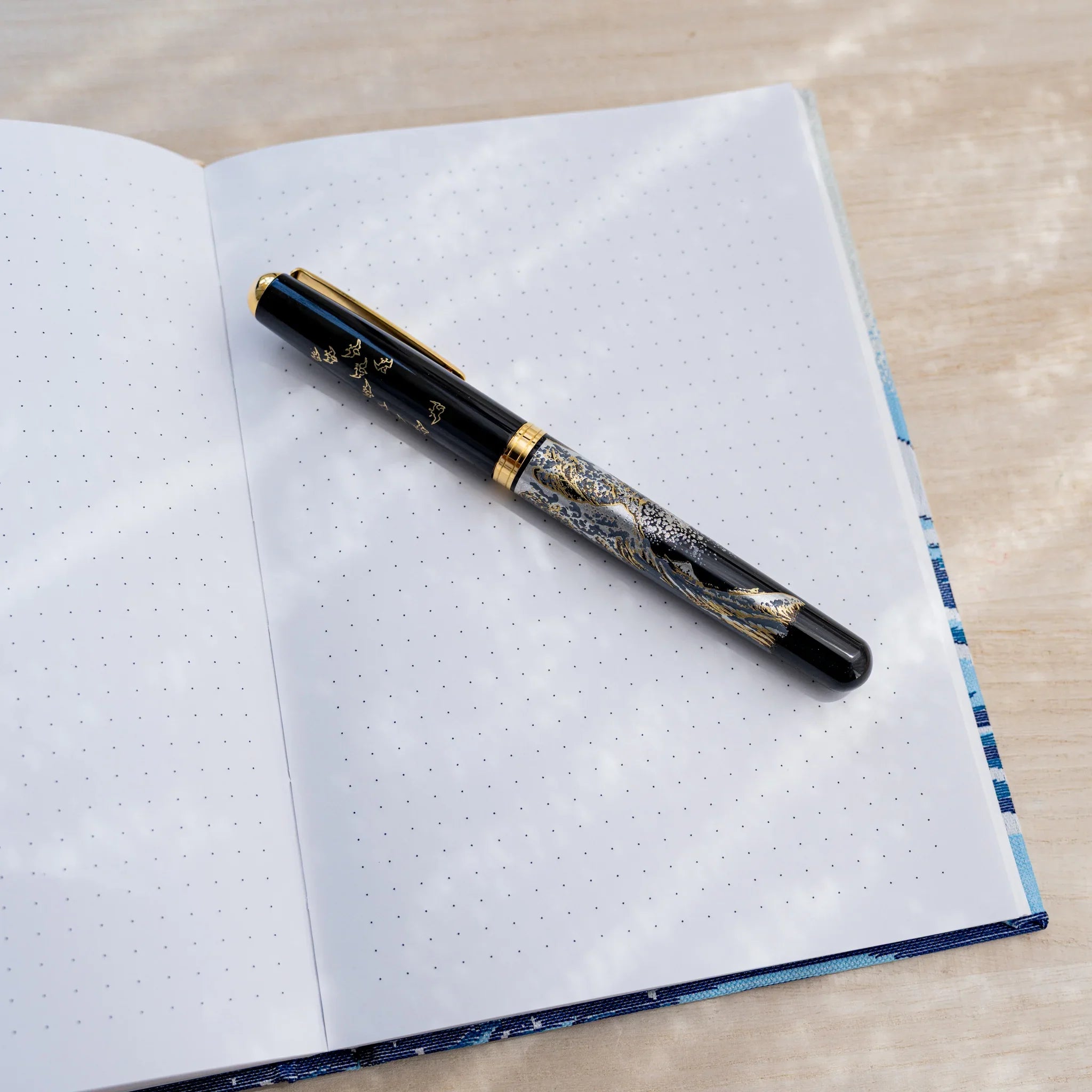 Notebook open to dot grid pages with a black fountain pen resting diagonally, blue textile cover edge