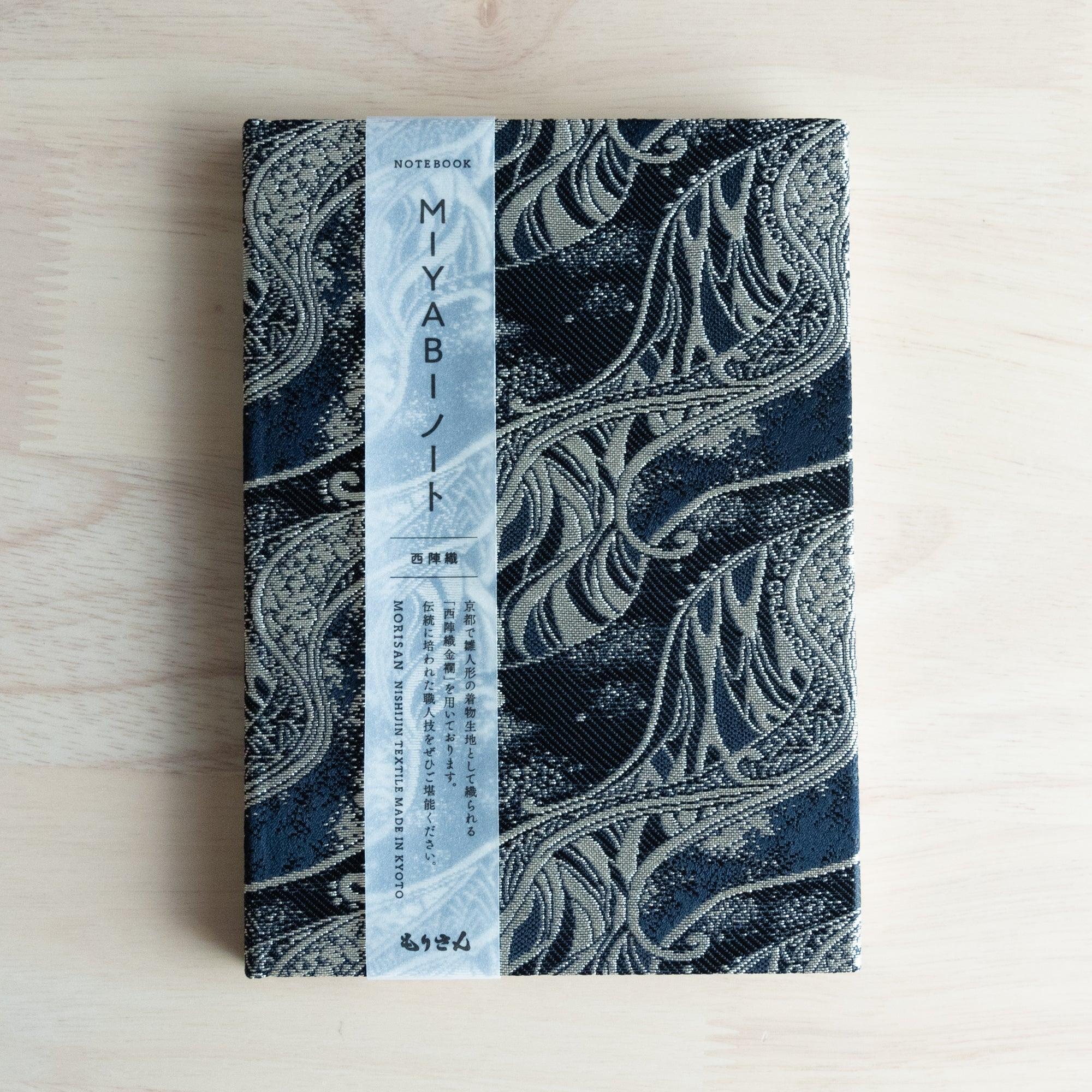A Japanese notebook with an elegant black fabric cover featuring intricate silver wave-like patterns, placed on a wooden table.