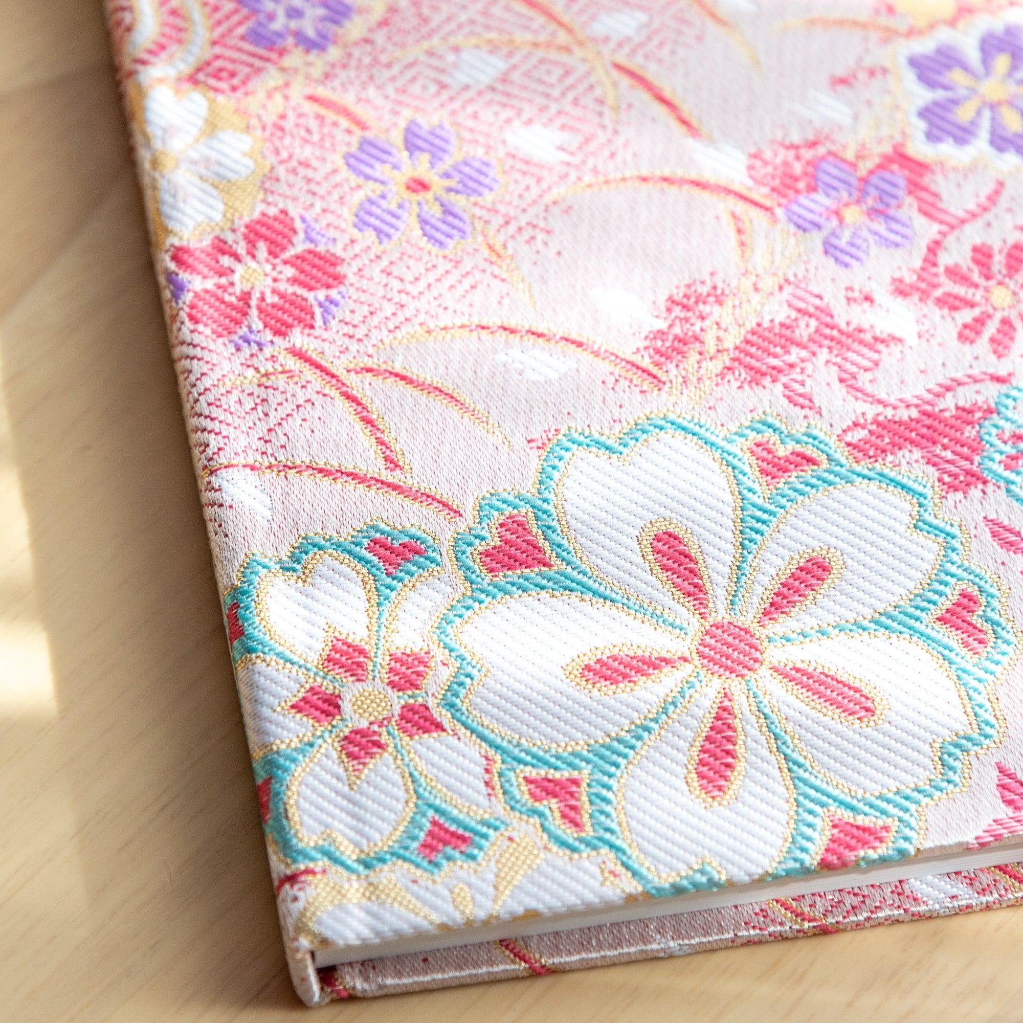 A Japanese notebook with a fabric cover featuring vibrant floral patterns in red, pink, and gold tones, placed on a wooden table.