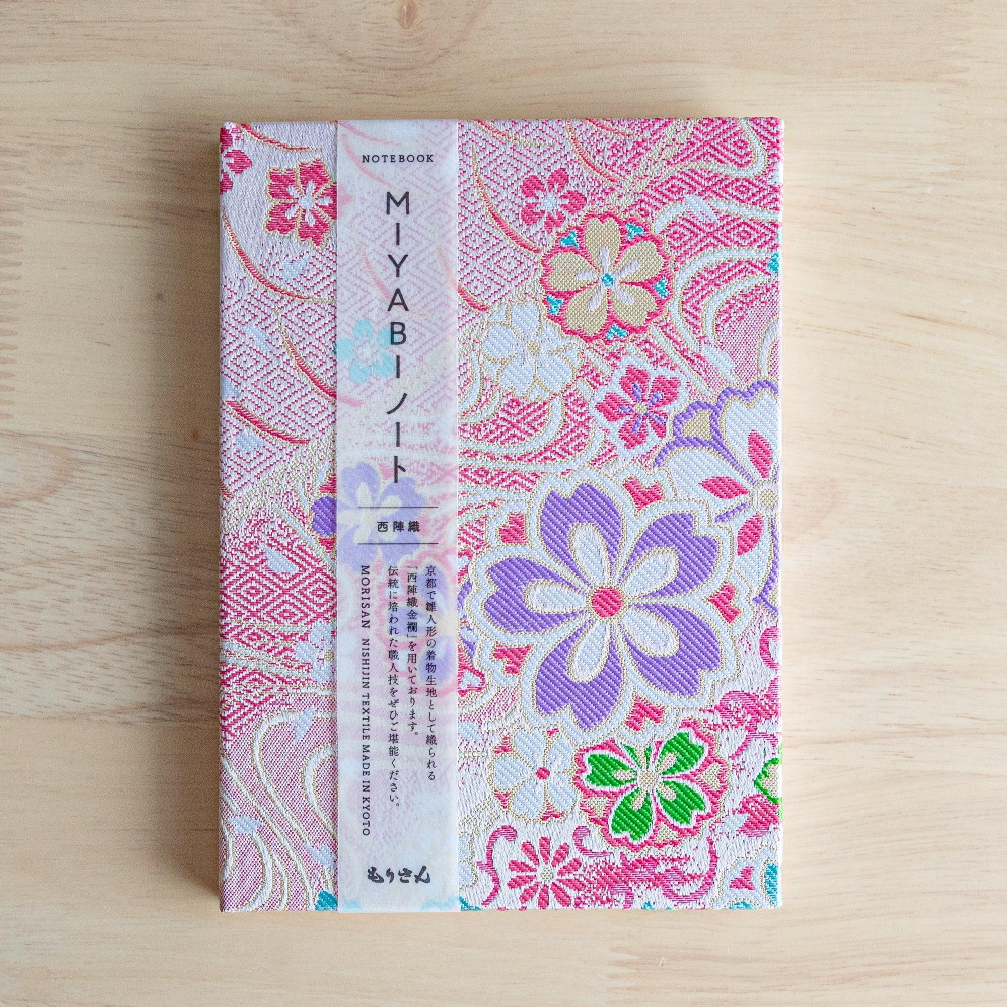 A Japanese notebook with a fabric cover featuring vibrant floral patterns in red, pink, and gold tones, placed on a wooden table.