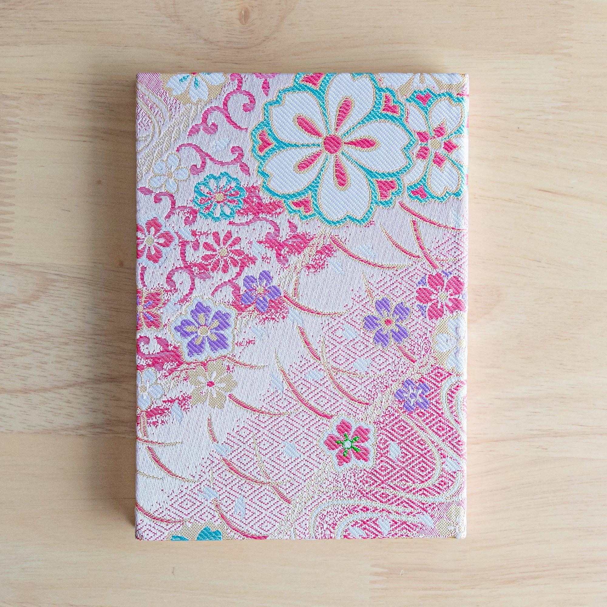 A Japanese notebook with a fabric cover featuring vibrant floral patterns in red, pink, and gold tones, placed on a wooden table.