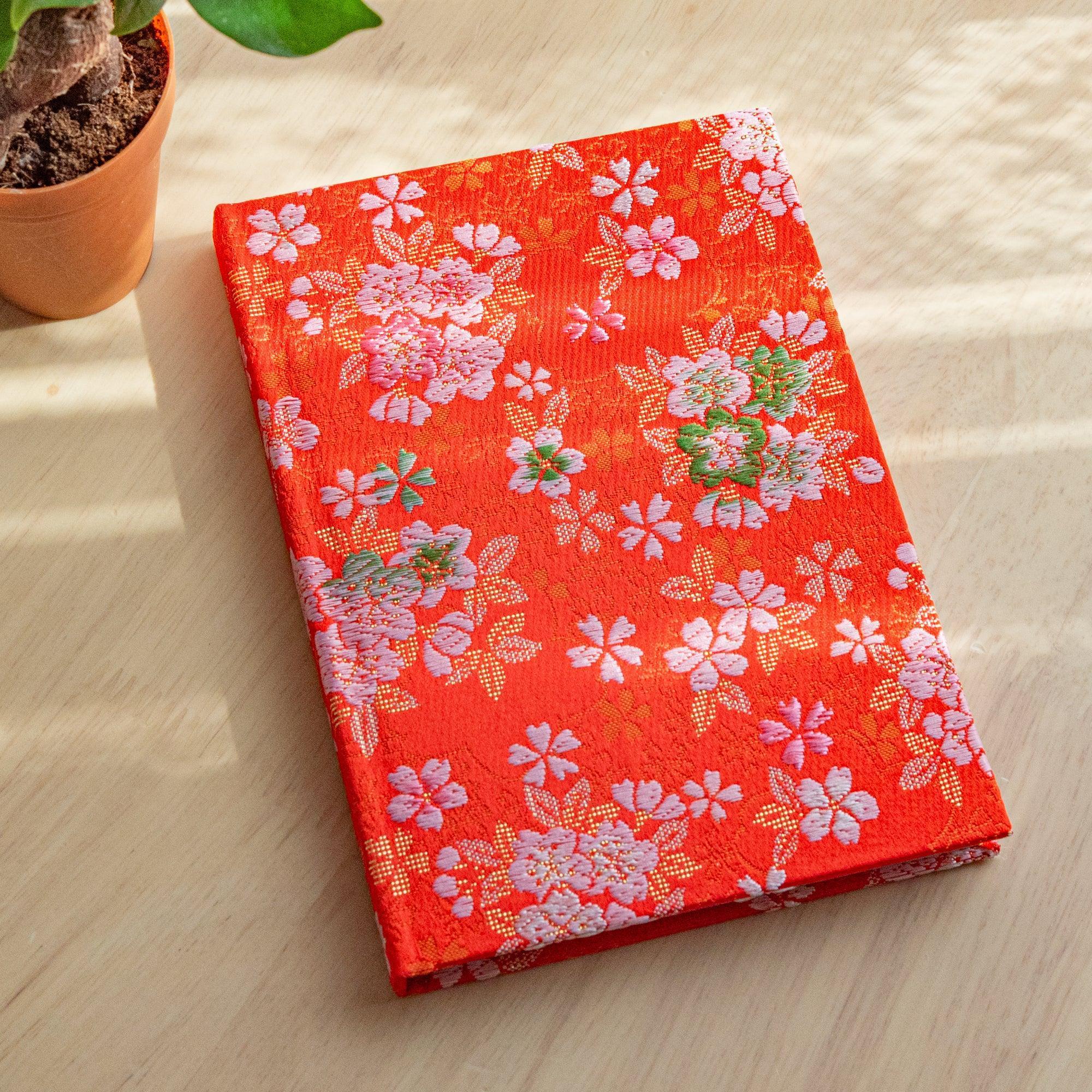 A Japanese notebook with a vibrant red fabric cover adorned with pink and white cherry blossom patterns, placed on a wooden table.