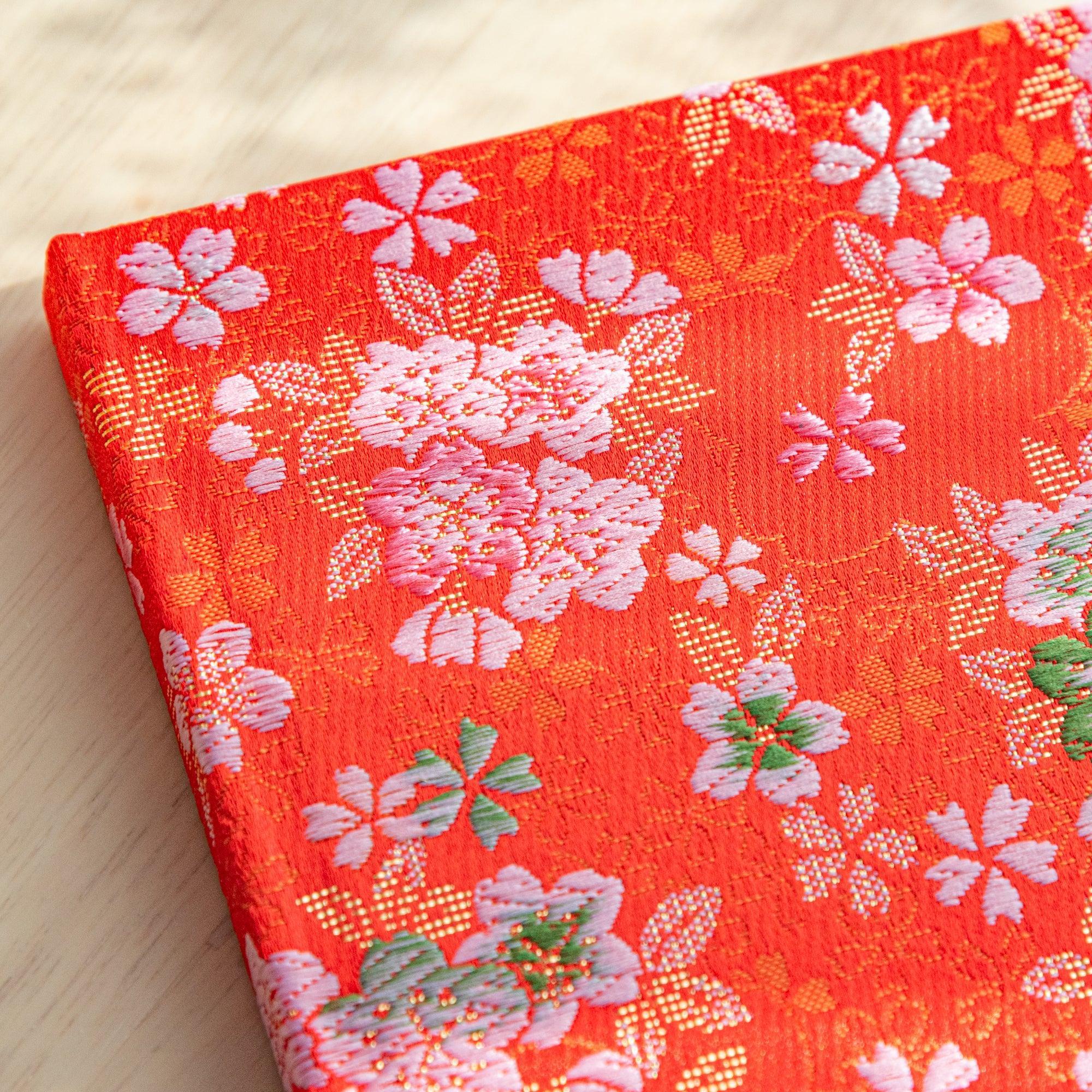 A Japanese notebook with a vibrant red fabric cover adorned with pink and white cherry blossom patterns, placed on a wooden table.