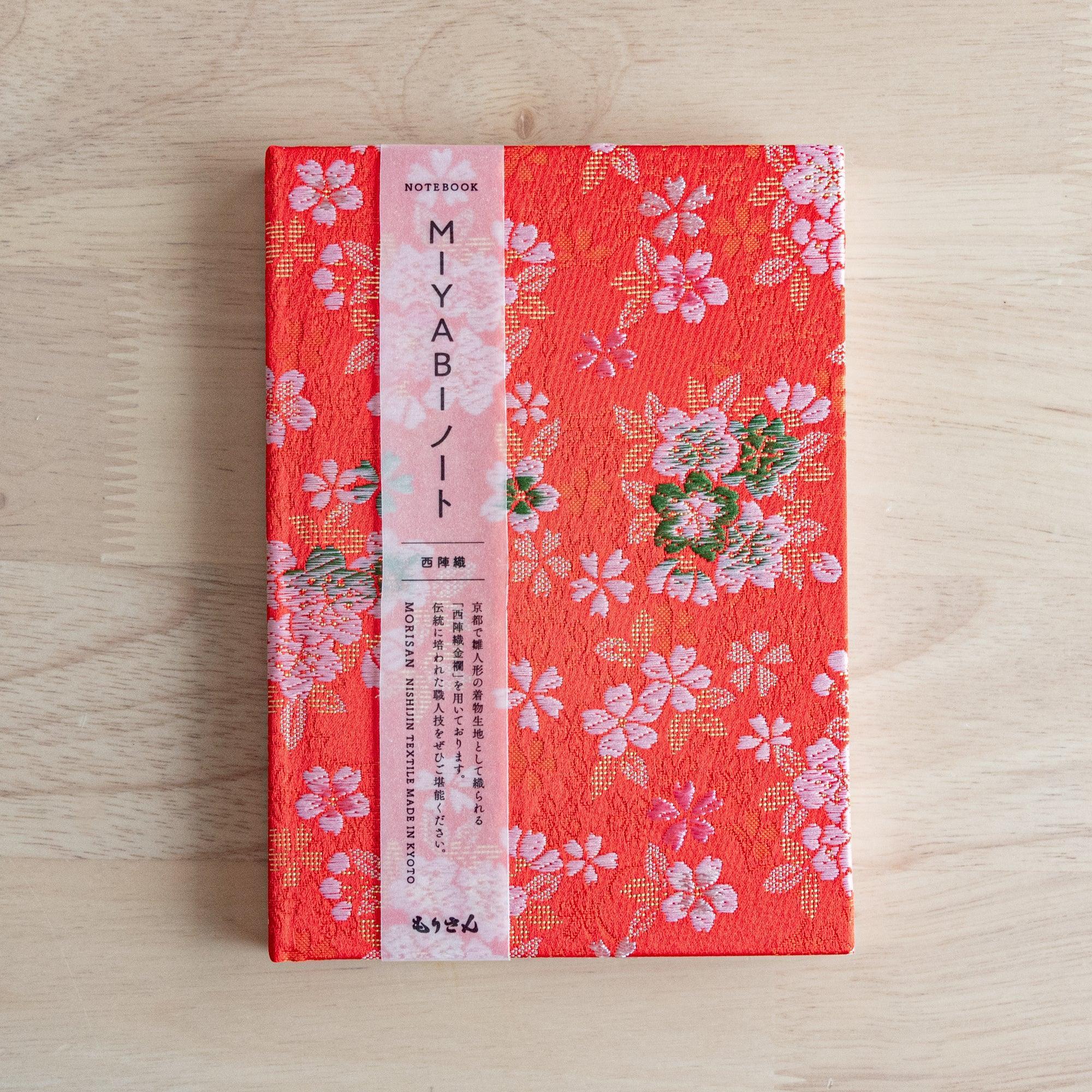 A Japanese notebook with a vibrant red fabric cover adorned with pink and white cherry blossom patterns, placed on a wooden table.