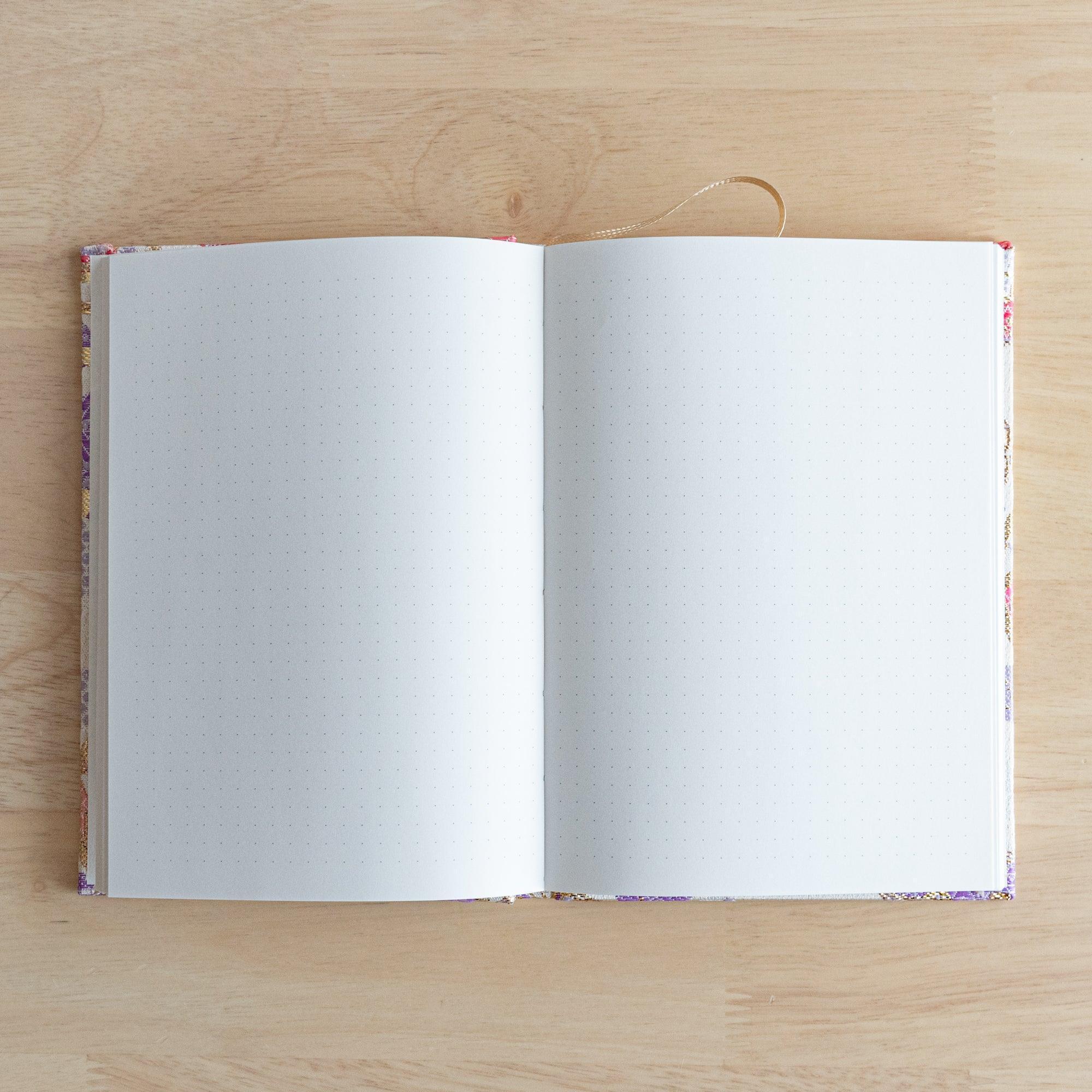 An open Japanese notebook showing white pages with a subtle dot grid pattern and a gold ribbon bookmark on a wooden table.