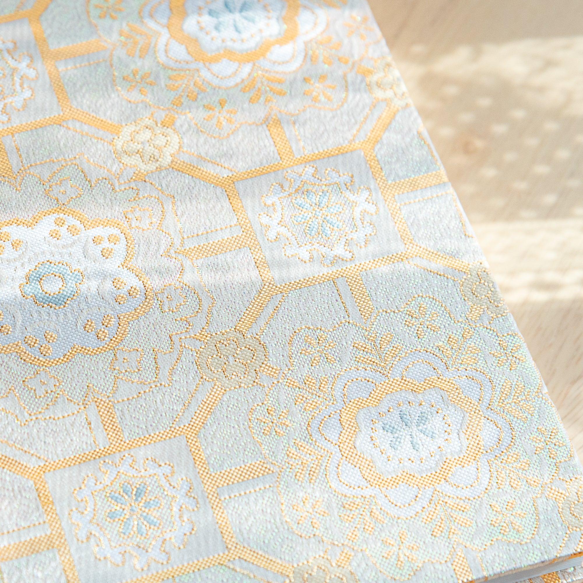 A Japanese notebook with a fabric cover featuring intricate gold and white patterns inspired by traditional motifs, placed on a wooden table.