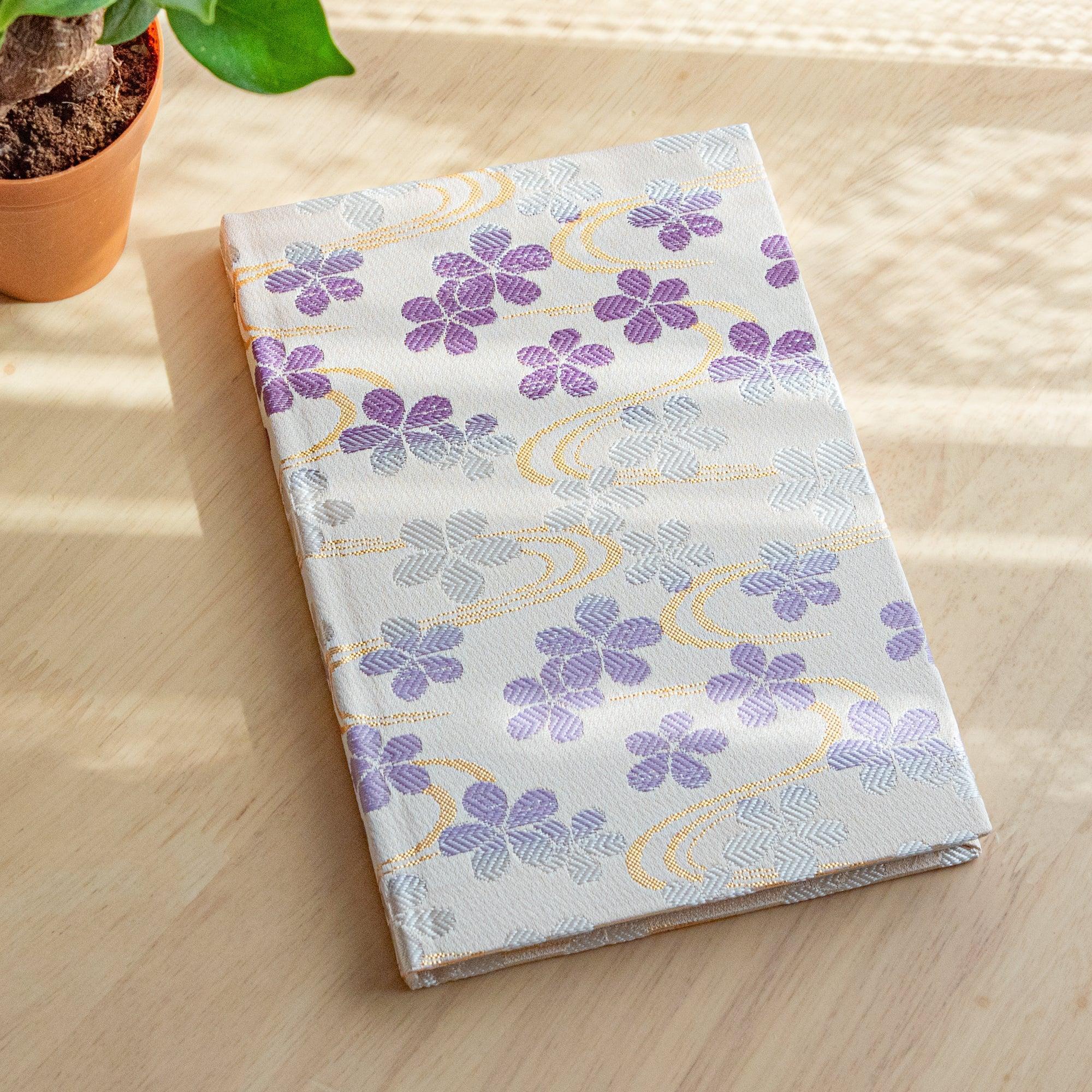 A Japanese notebook with a white fabric cover featuring delicate purple floral patterns and flowing gold accents, placed on a wooden table.