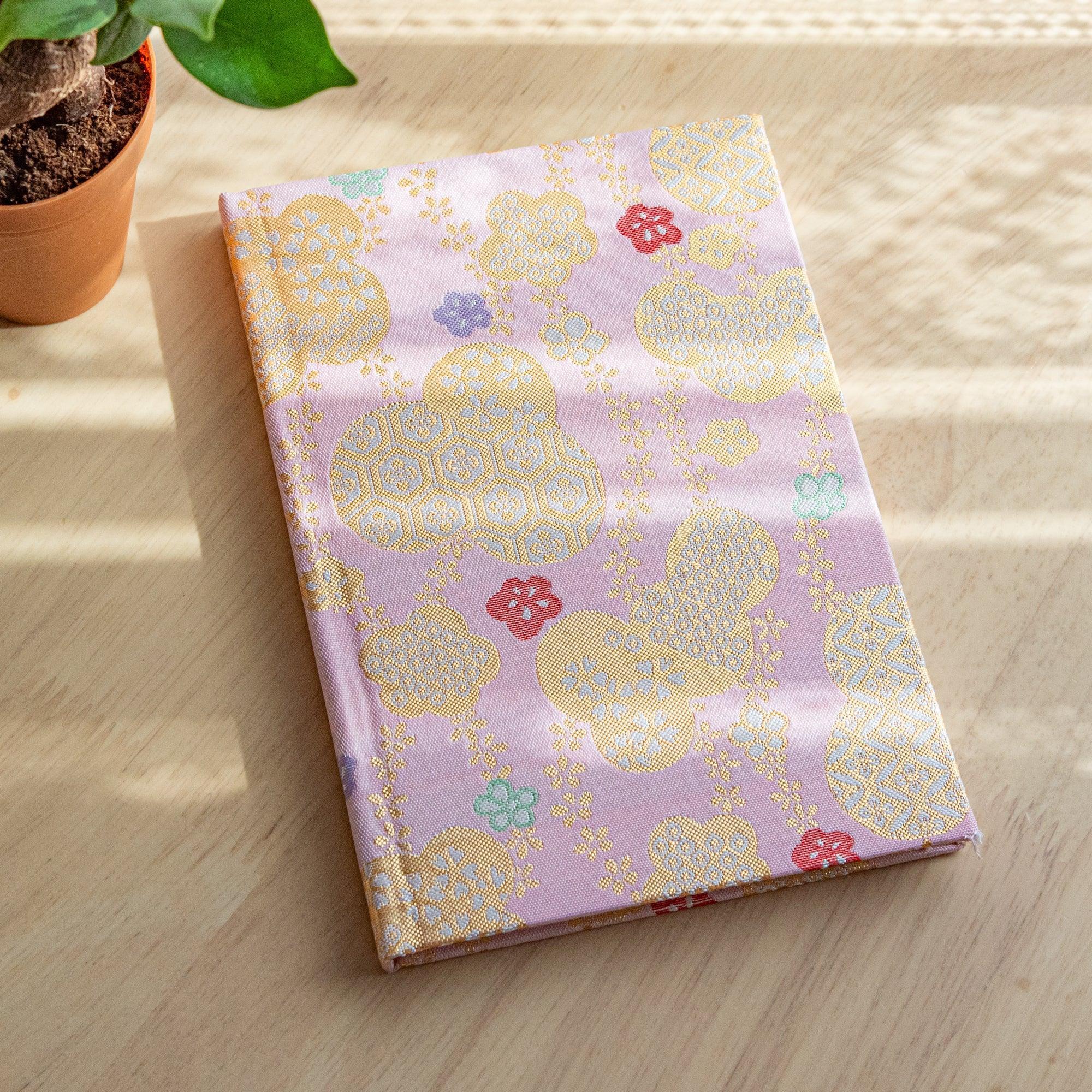 A Japanese notebook with a pink fabric cover featuring traditional patterns in gold and pastel tones, placed on a wooden table.