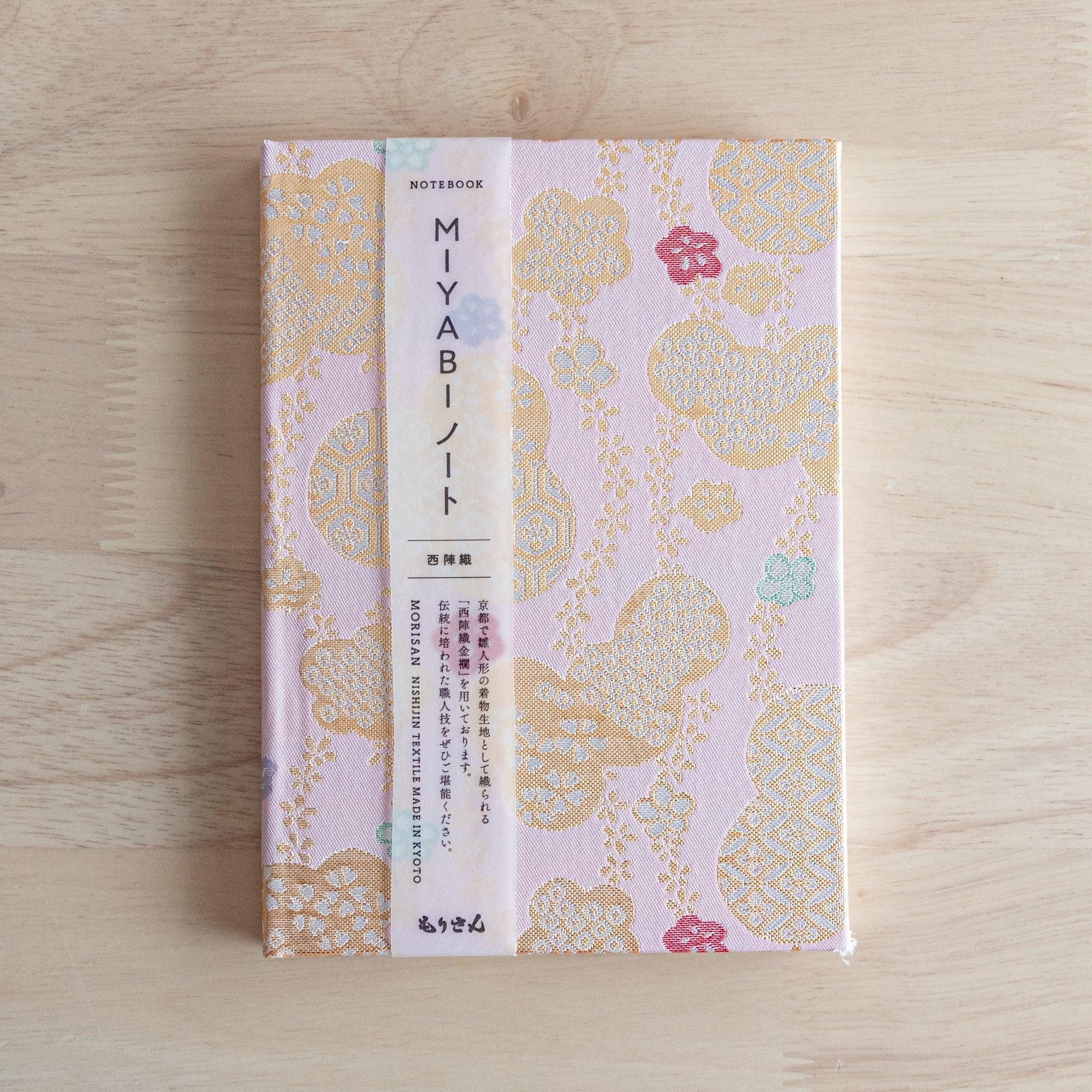 The front view of a "MIYABI" Japanese notebook with a pink fabric cover and a product label describing the craftsmanship.