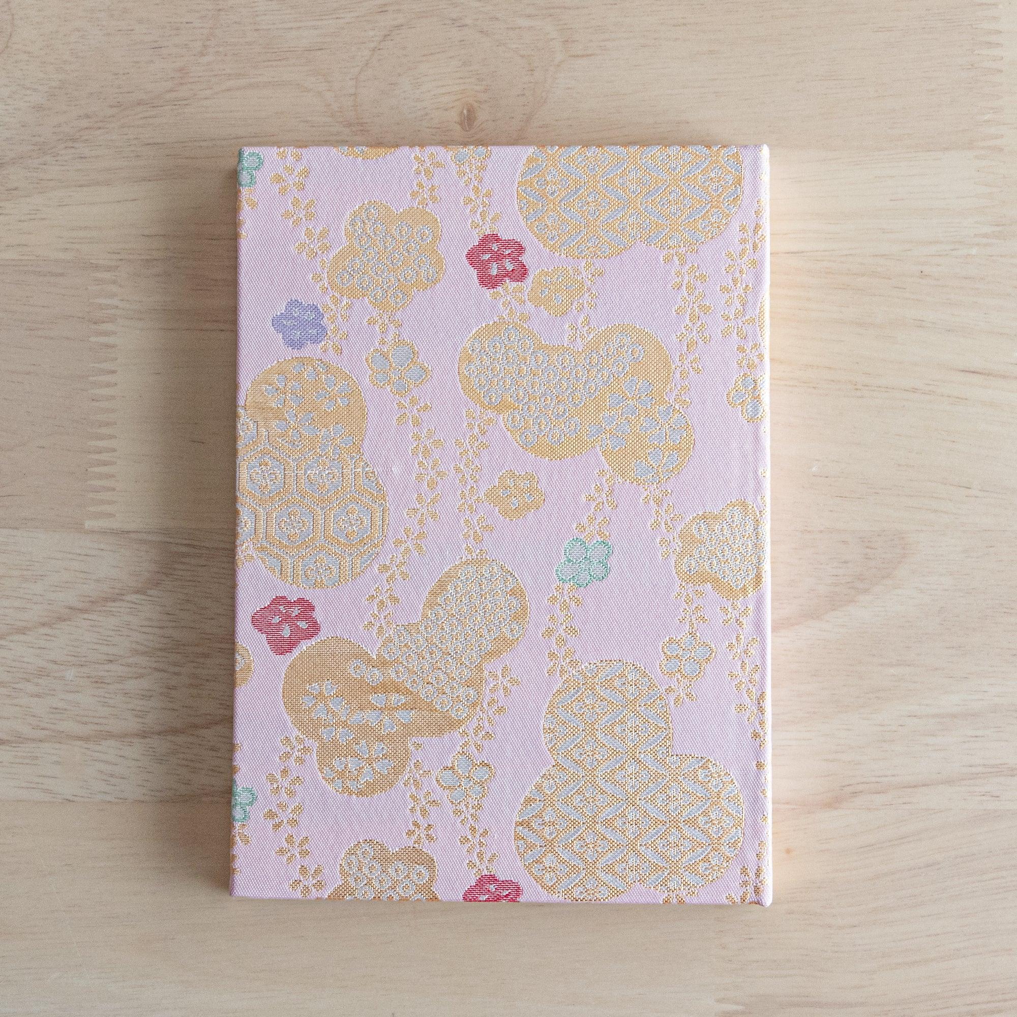The back view of a Japanese notebook with a pink fabric cover decorated with gold and pastel cloud and floral patterns.