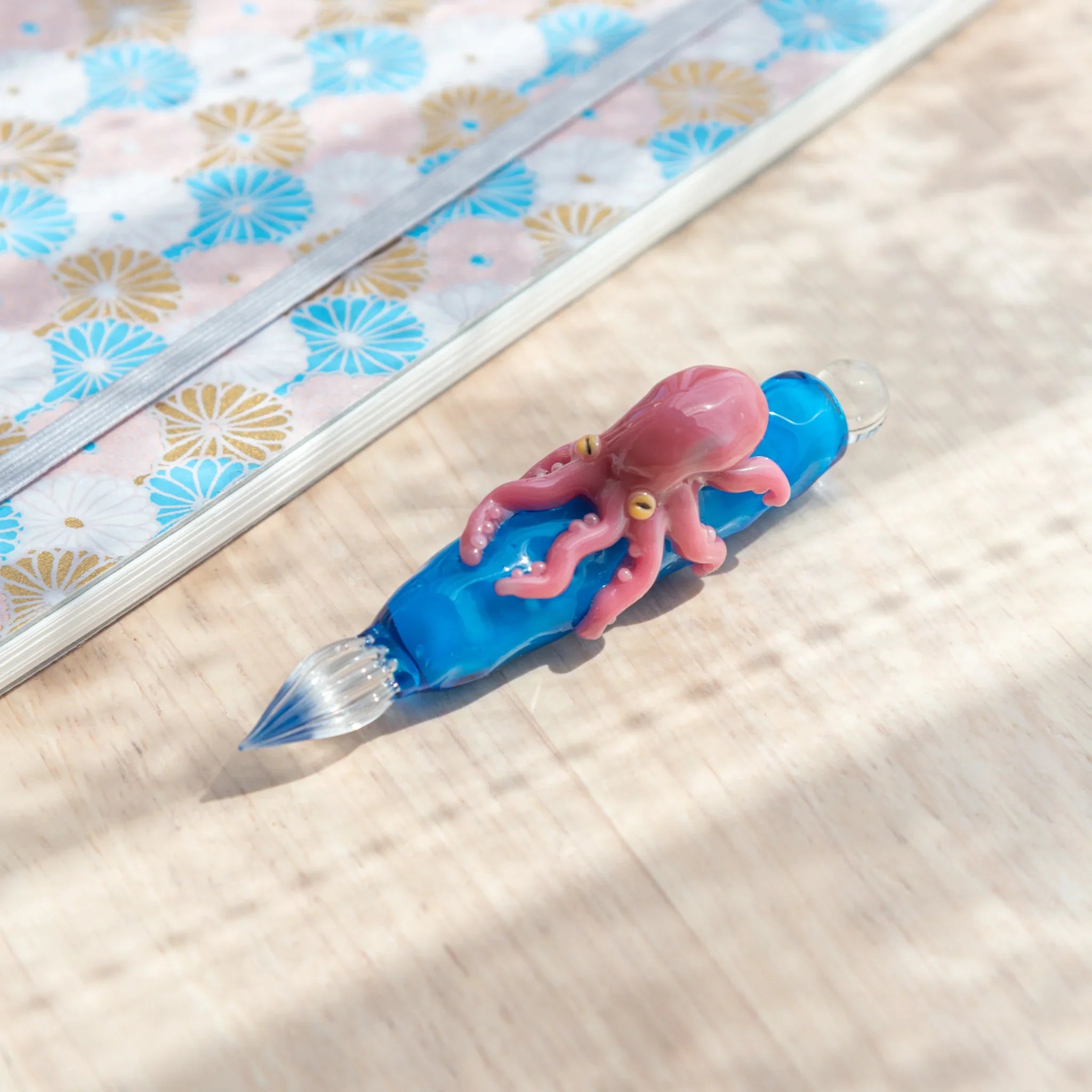 Blue glass Japanese dip pen with pink octopus figurine lying diagonally on light wood surface beside a blue floral notebook