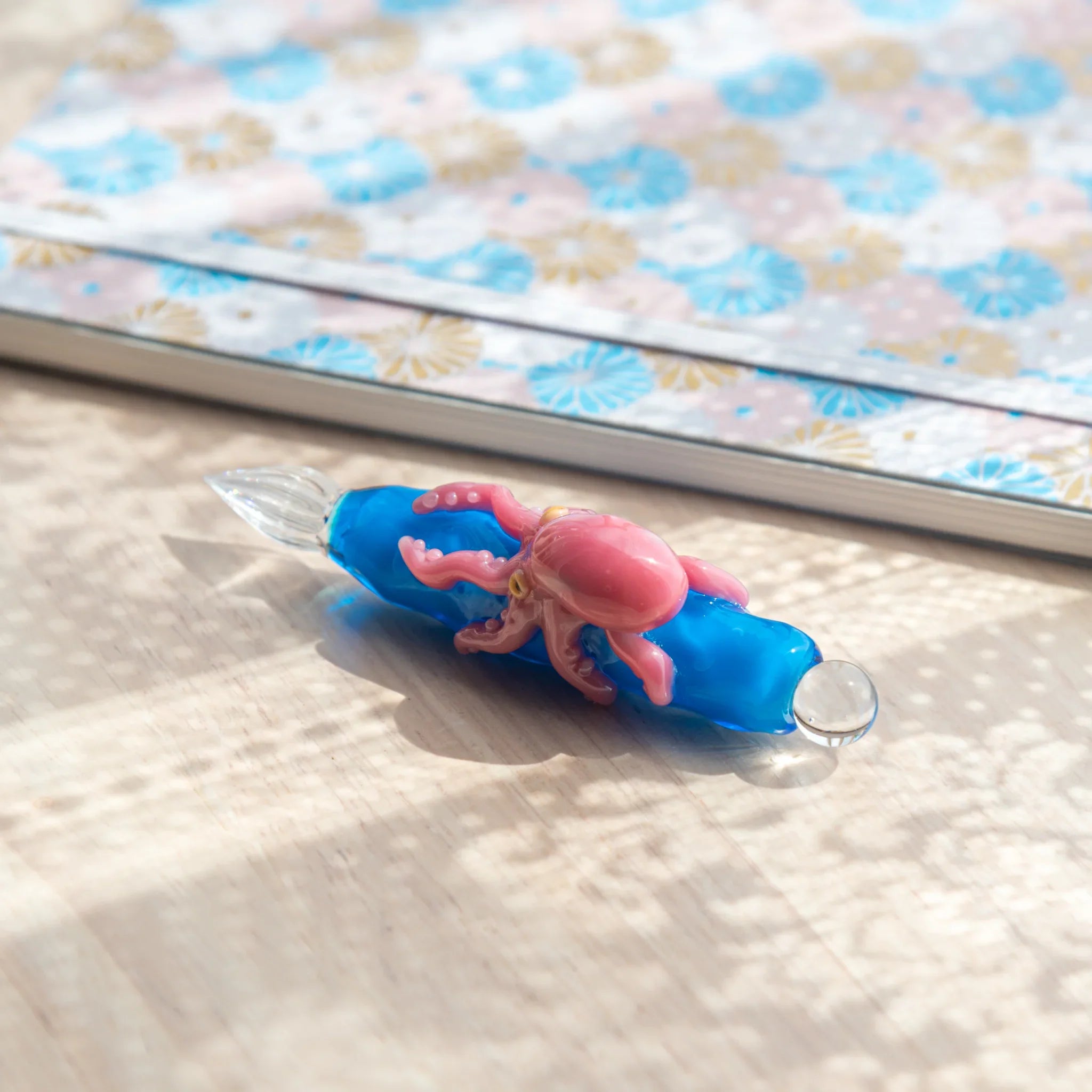 Full view of blue glass Japanese dip pen with pink octopus figurine, clear nib and ball cap visible, on light wood surface