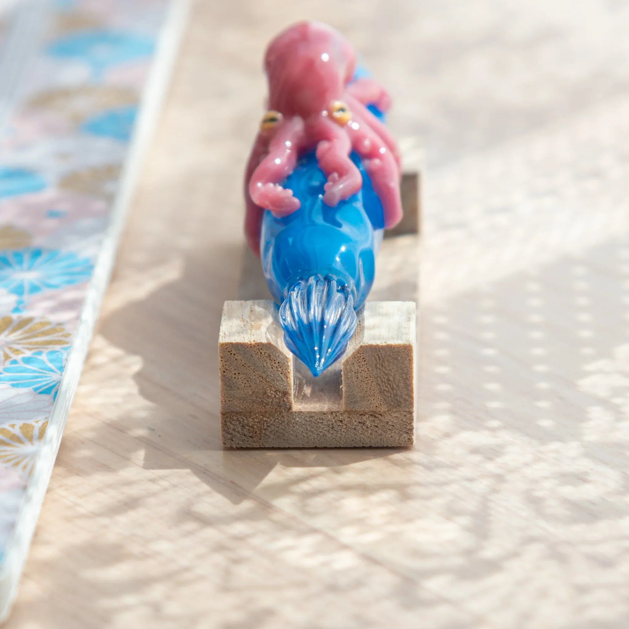 Blue glass Japanese dip pen with pink octopus on natural wood pen rest, clear glass nib in groove, viewed from the front