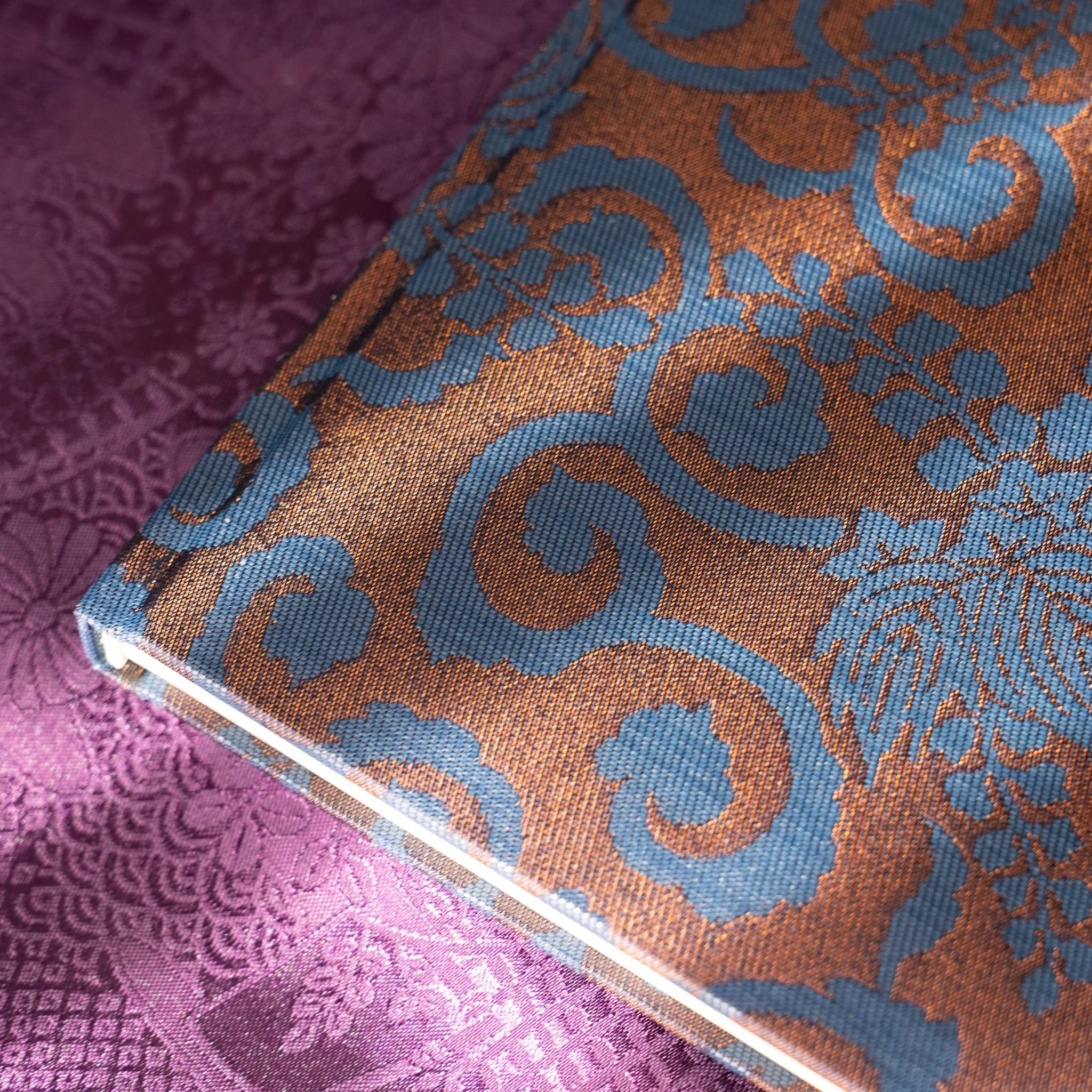 Oni Kiri Nishijin silk cover lower corner macro showing blue arabesque scroll pattern on copper-brown Nishijin weave