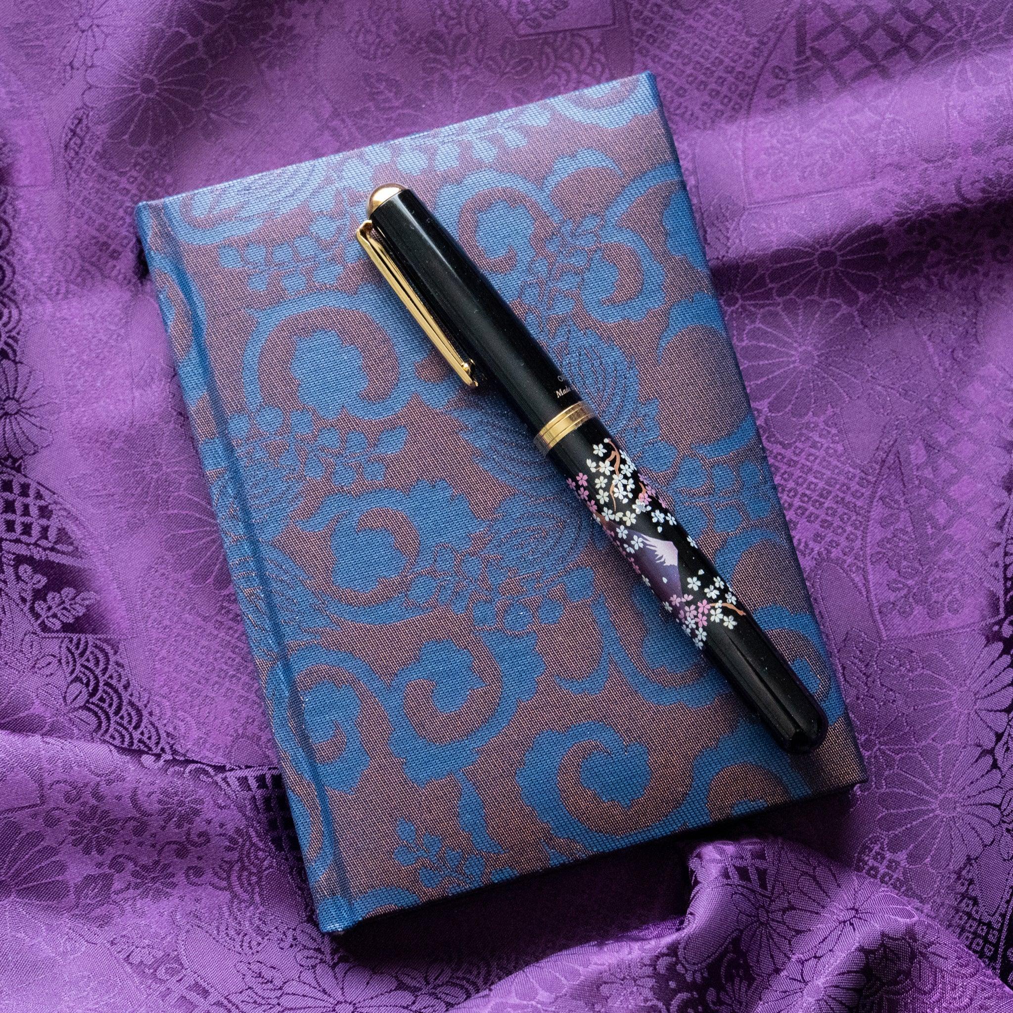 Oni Kiri Nishijin silk Homeicho notebook on purple fabric with black gold sakura maki-e fountain pen placed across the cover
