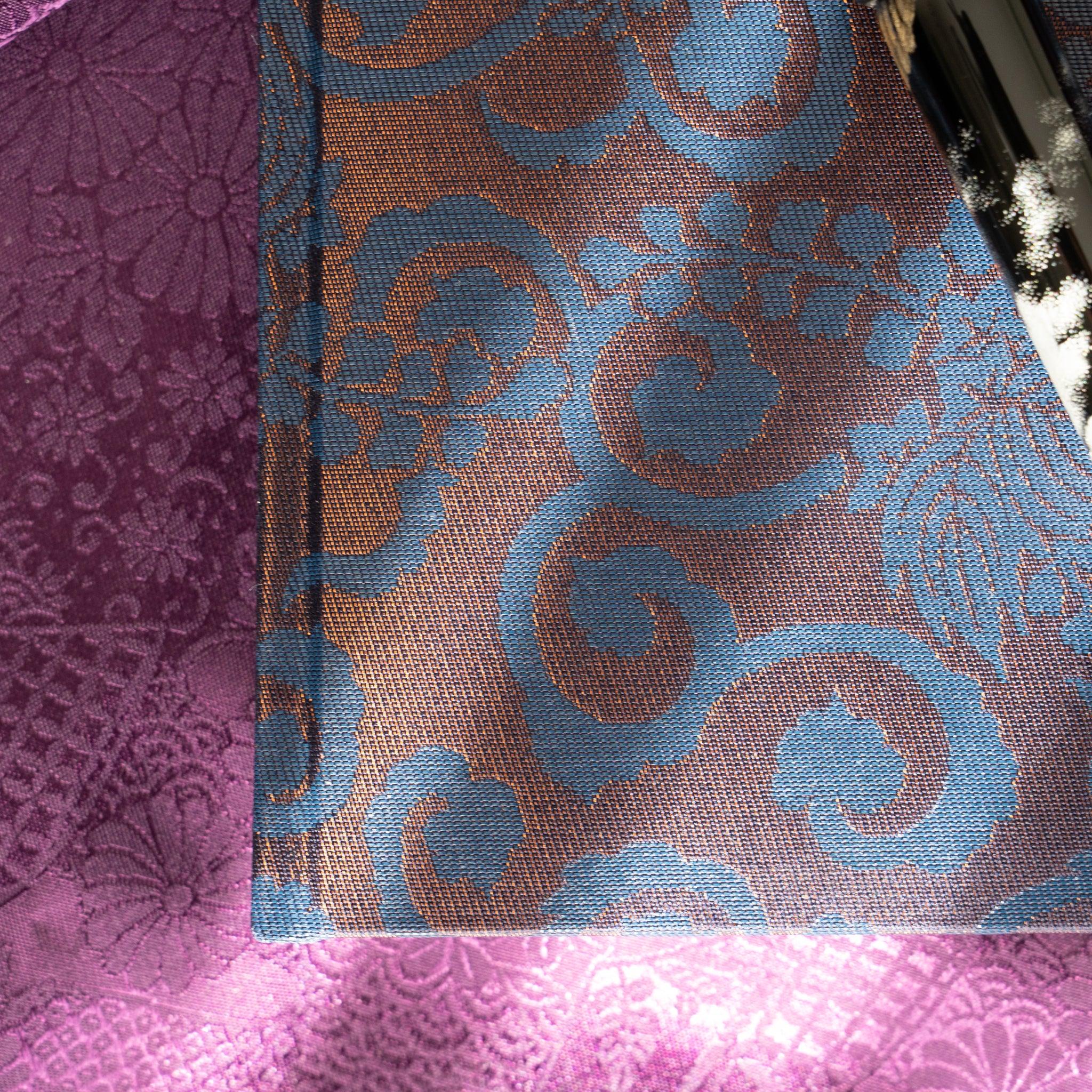 Angled close-up of Oni Kiri Nishijin silk cover showing blue scroll arabesque weave pattern on copper-toned background